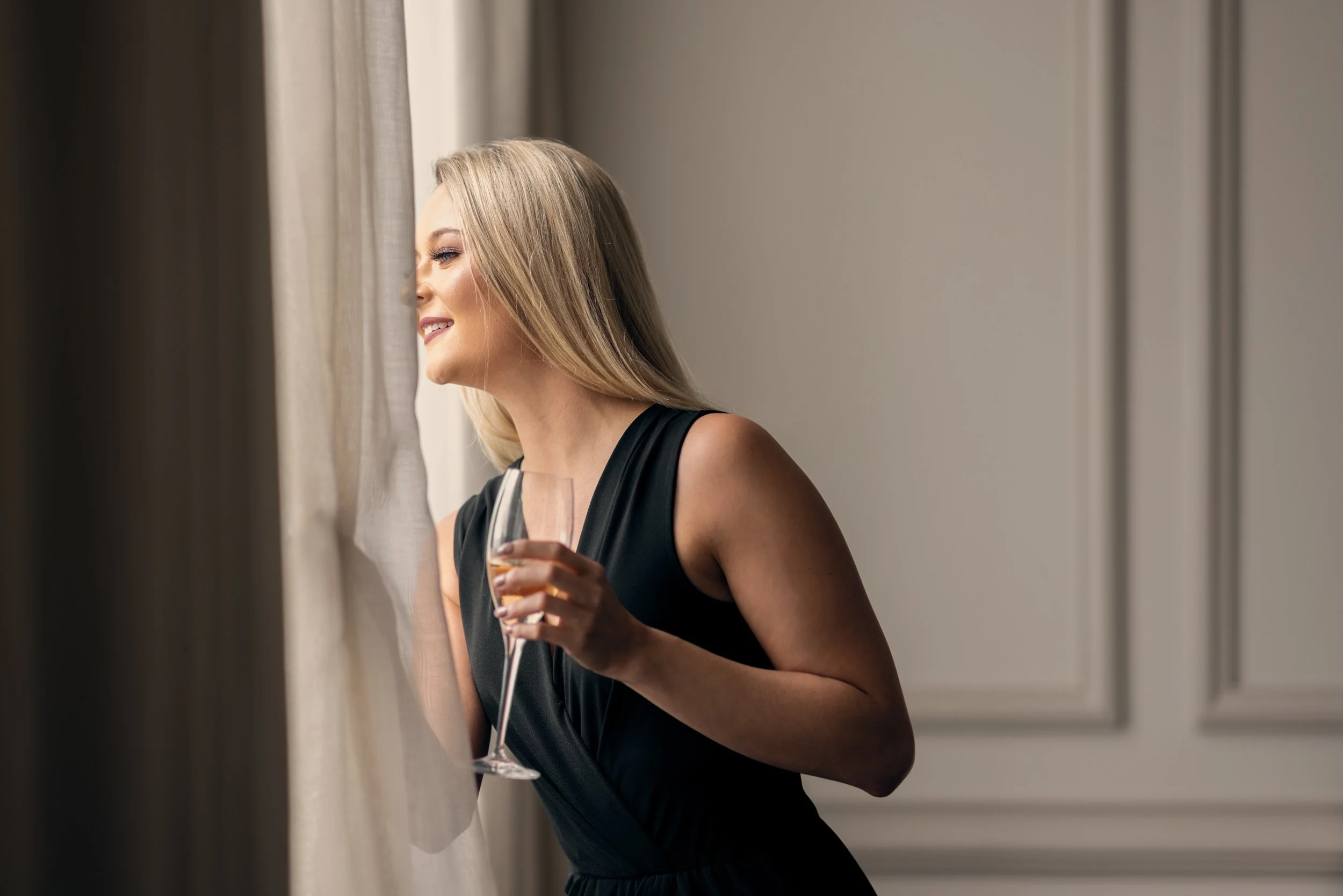 A woman in a black sleeveless dress with blonde hair is smiling as she looks out a window, holding a glass of wine.