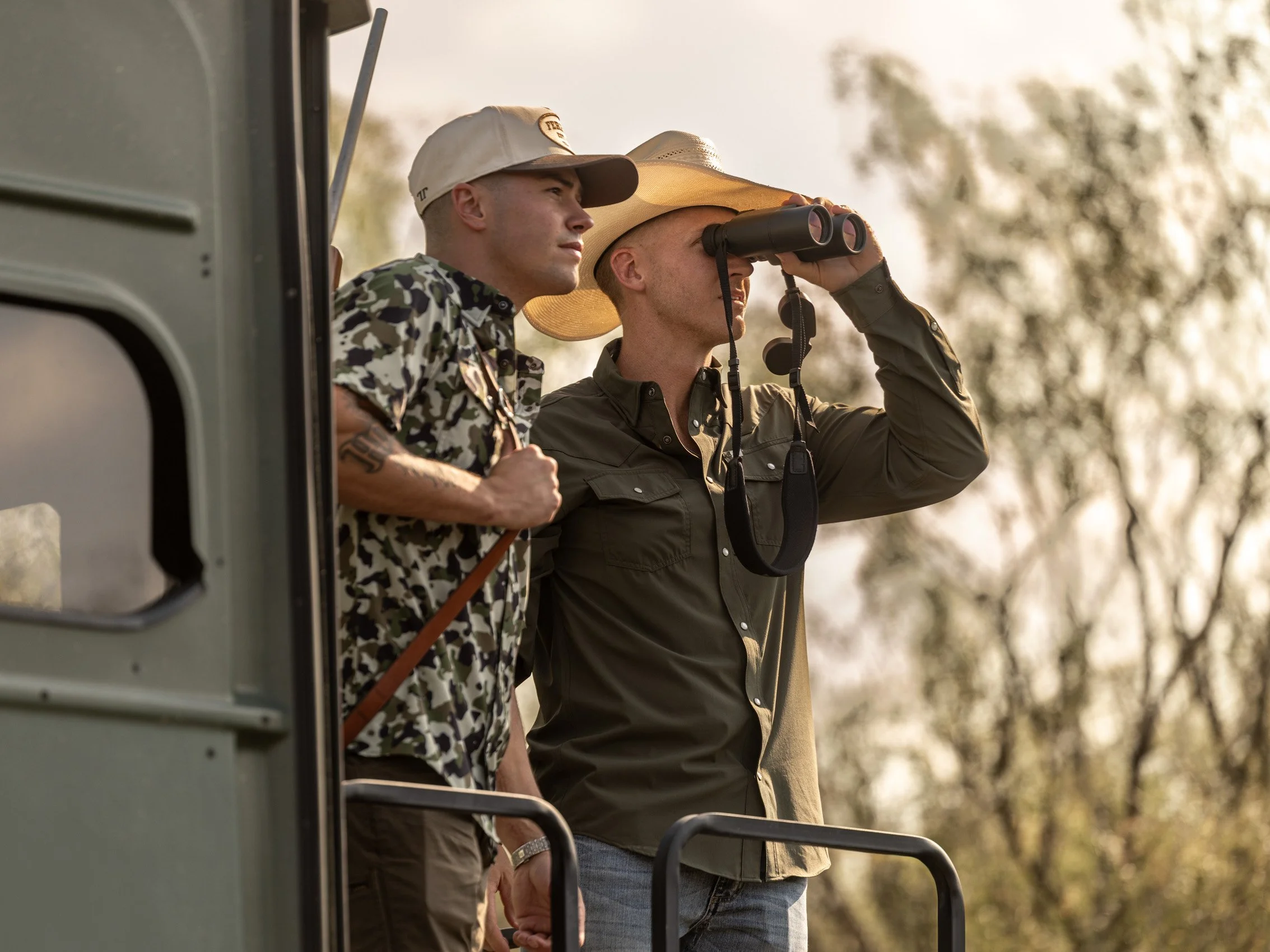 Two men are standing outdoors, looking into the distance. One holds binoculars, and the other is wearing a wide-brimmed hat. The image is taken near sunset or in warm lighting.