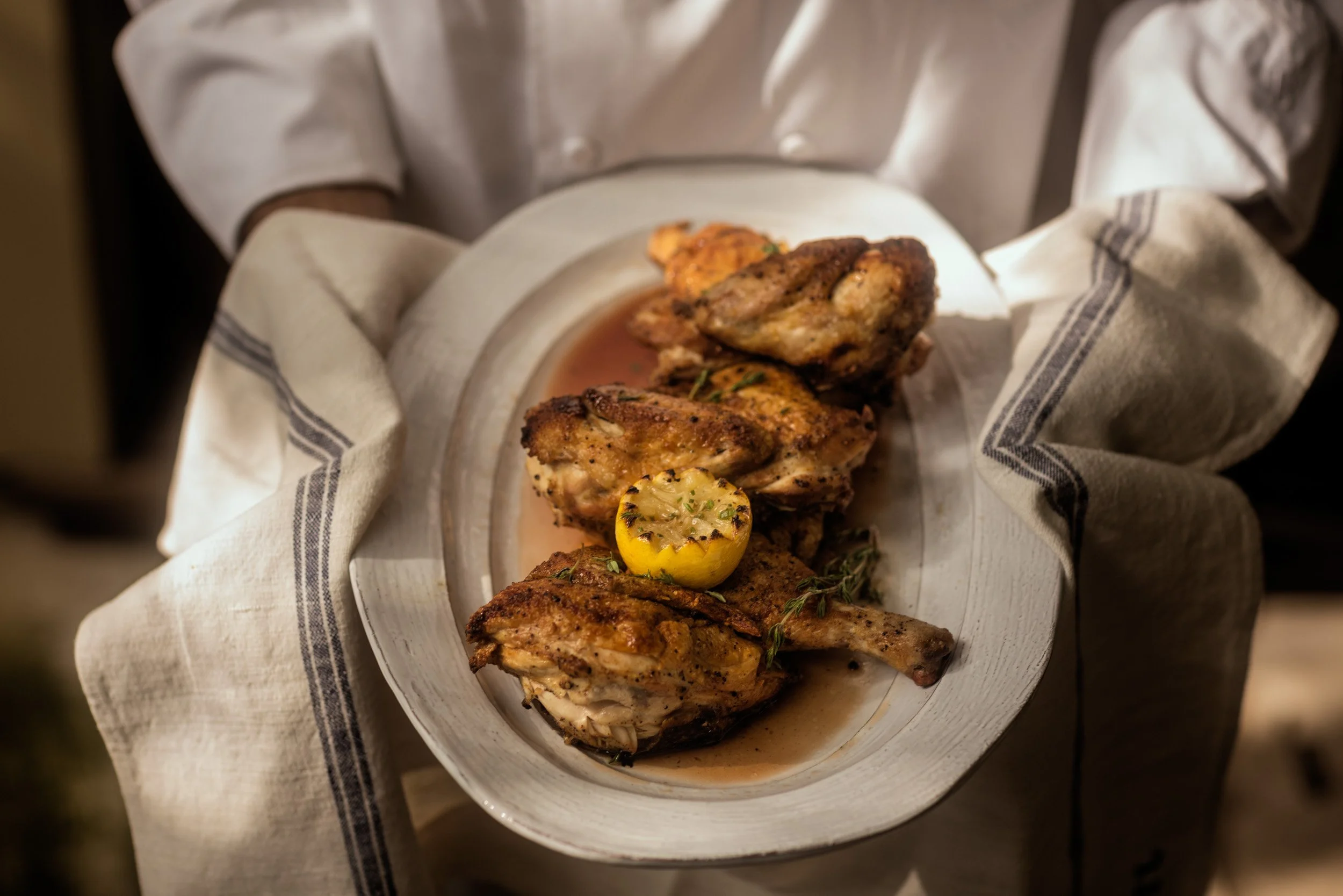 A white oval dish with grilled chicken pieces, garnished with herbs and a grilled lemon half, placed on a white cloth with black stripes.