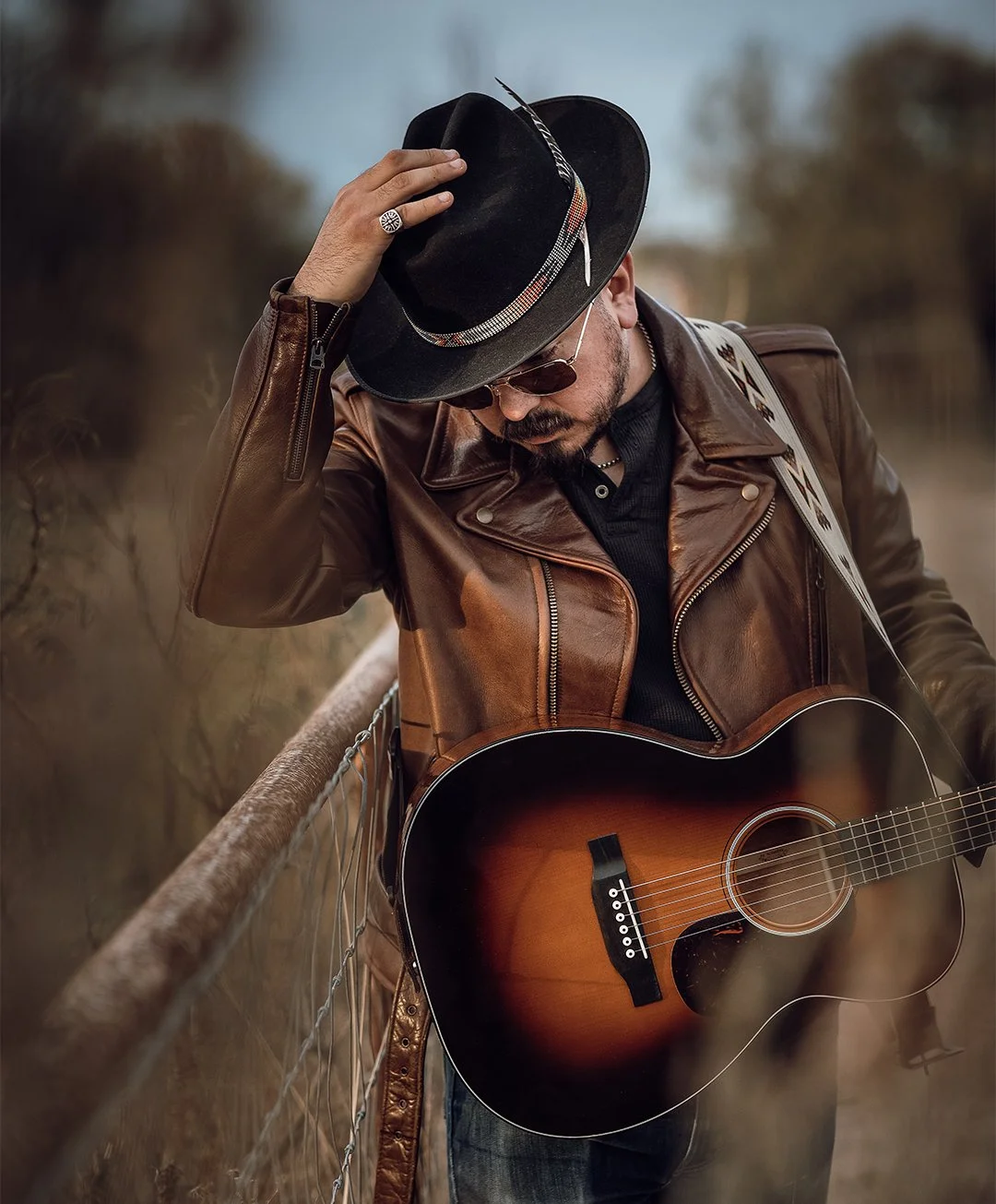 A man wearing a brown leather jacket, black shirt, sunglasses, and a black hat with a feather, playing an acoustic guitar outdoors near a fence with a blurred background.