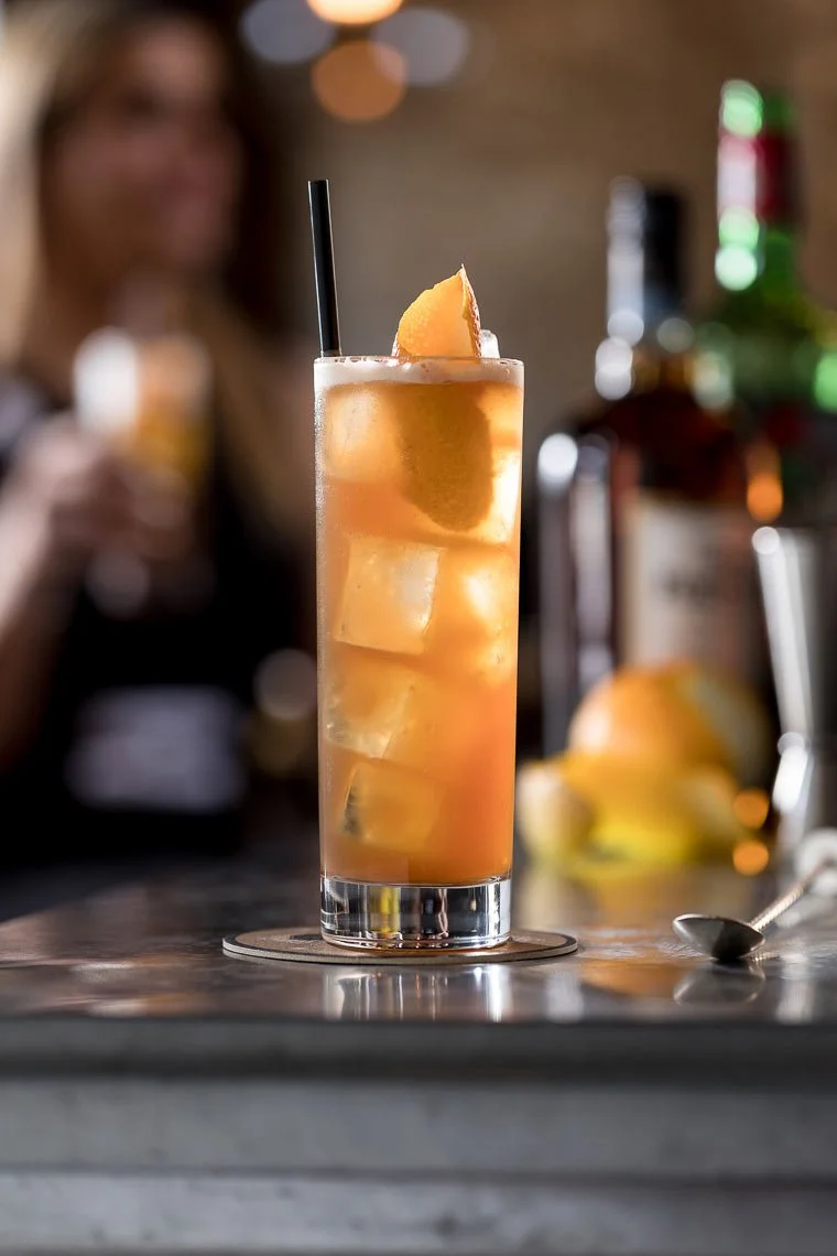 A tall glass of iced peach tea with a lemon wedge garnish and a black straw on a bar counter, blurred woman in background holding a drink.