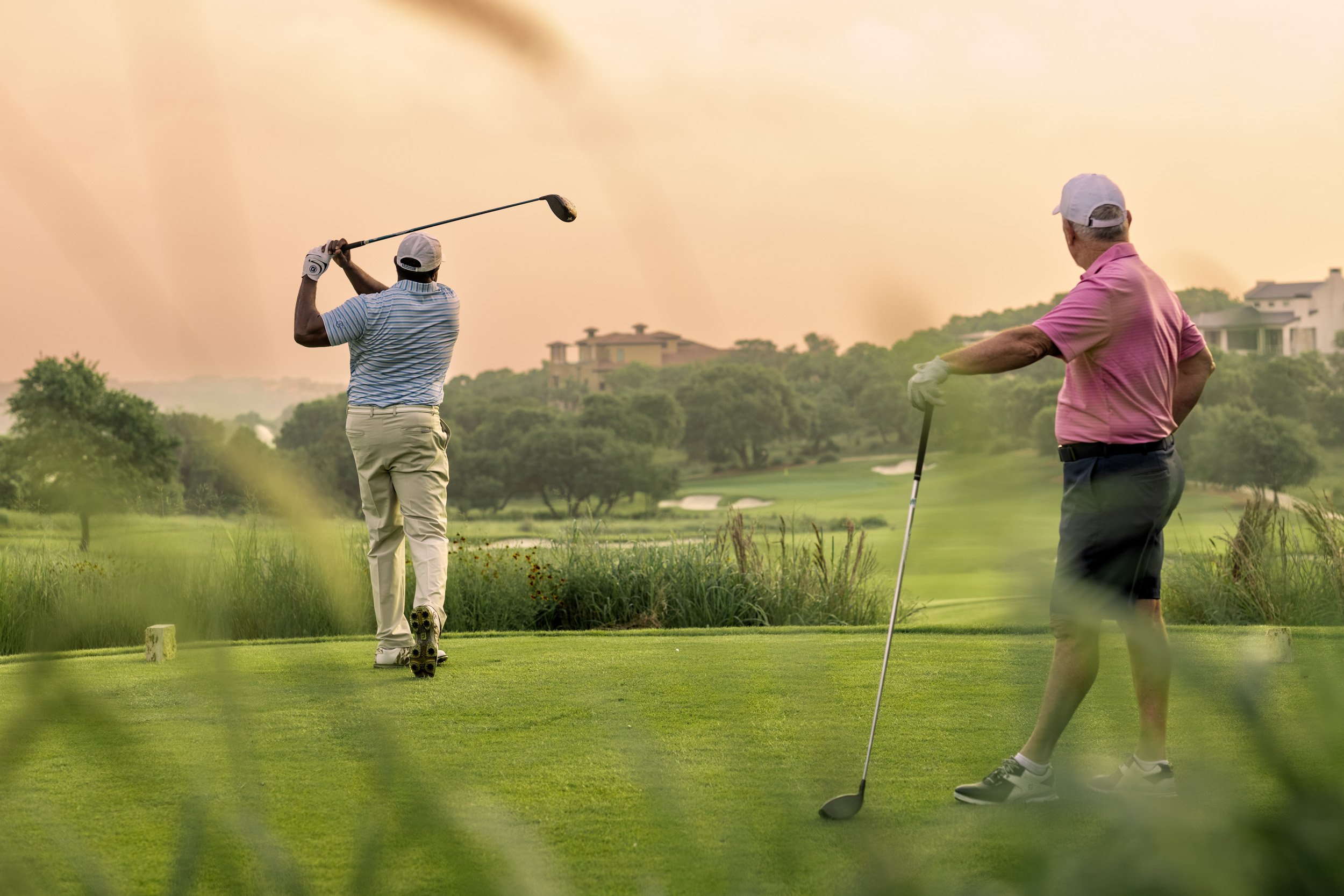 Two men playing golf on a lush golf course during sunset. One man is swinging a golf club, and the other man is standing with a golf club. There are houses in the background and trees surrounding the course.