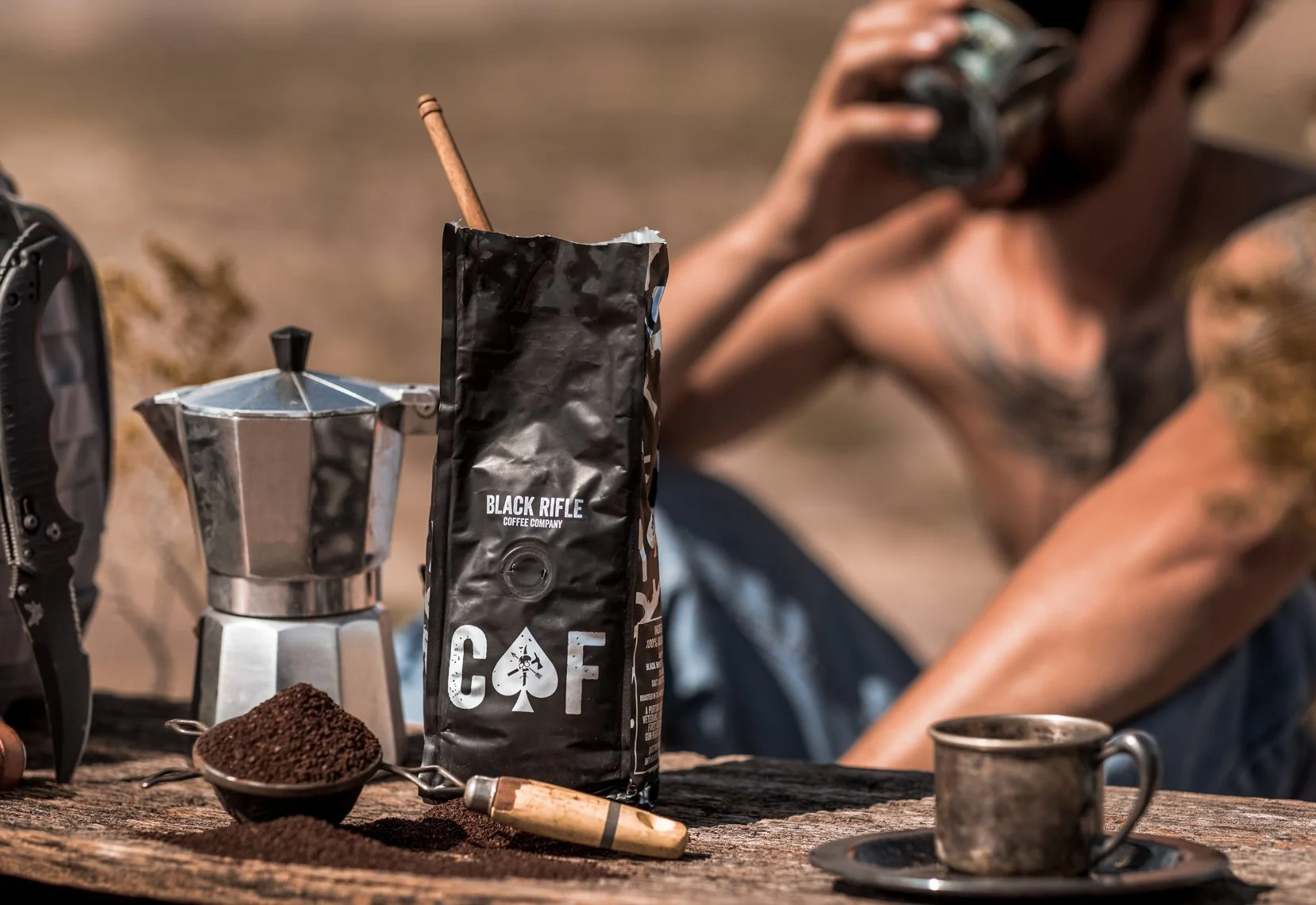 A person sitting outdoors drinking from a mug, with a bag of Black Rifle Coffee Company coffee and a moka pot coffee maker on a rustic wooden table, surrounded by coffee grounds and a spoon.