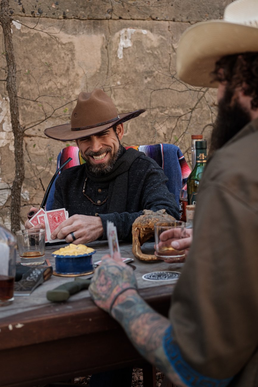 Two men playing cards at an outdoor table, smiling and enjoying drinks. One wears a large cowboy hat, and the other has a tattooed arm and a wide-brimmed hat. The setting appears rustic with a stone wall in the background.