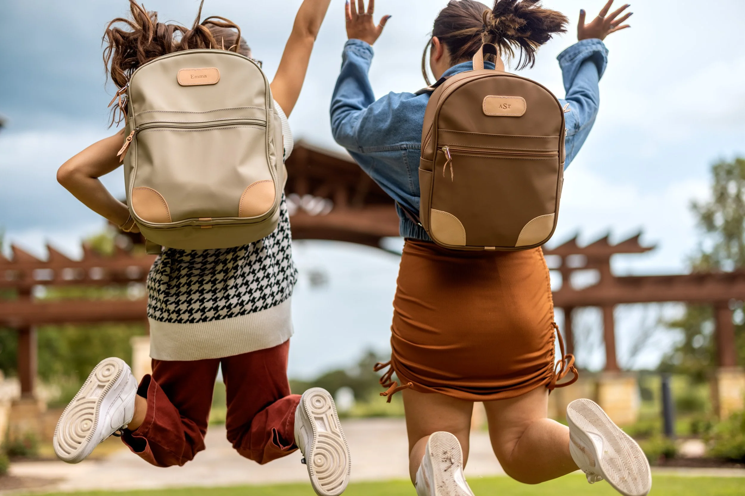 Two young women with backpacks jumping in a park during daytime.