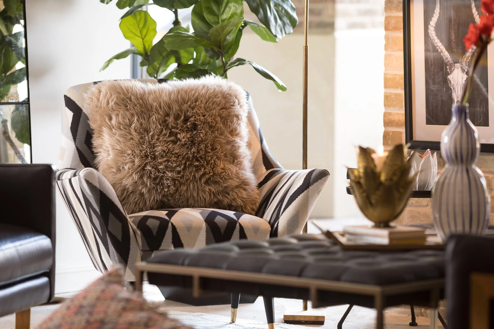 Living room with a patterned armchair holding a fluffy brown cushion, large green plant, framed artwork of a bull skull on brick wall, decorative vases, and a coffee table with a flower arrangement.