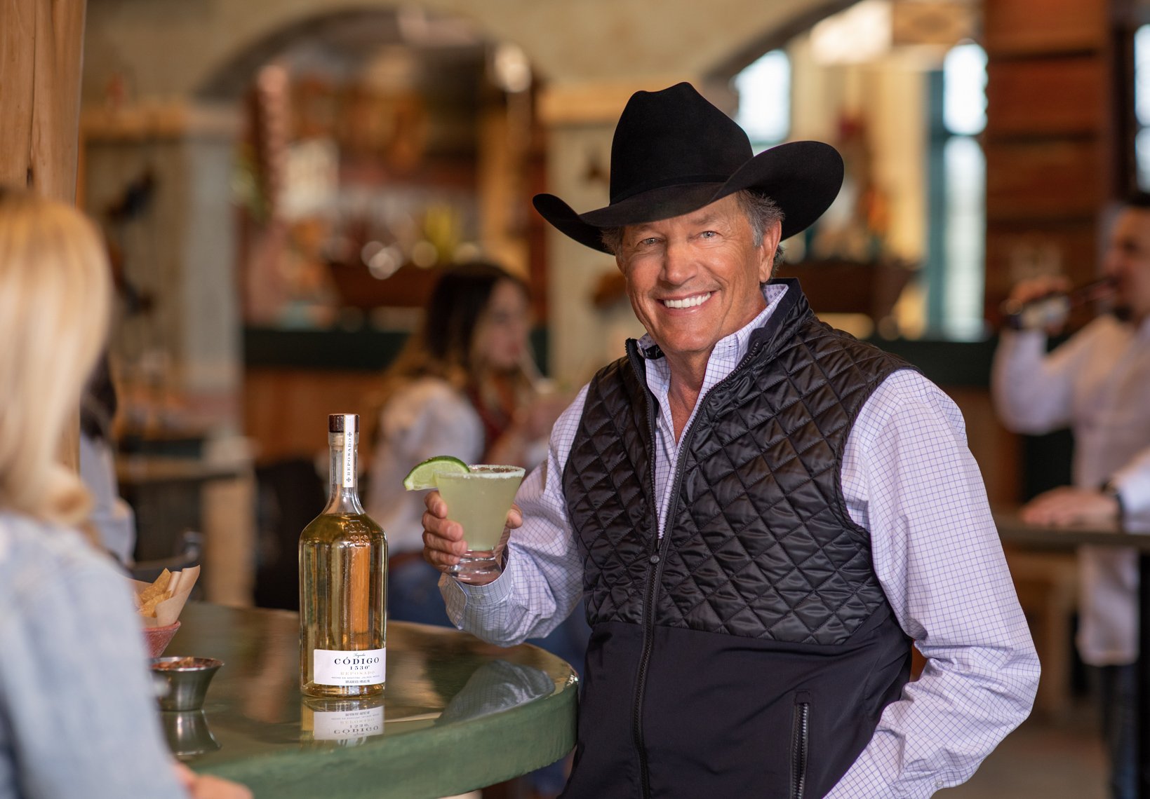 A smiling man in a cowboy hat and quilted vest holding a margarita with a lime wedge at a bar or restaurant.