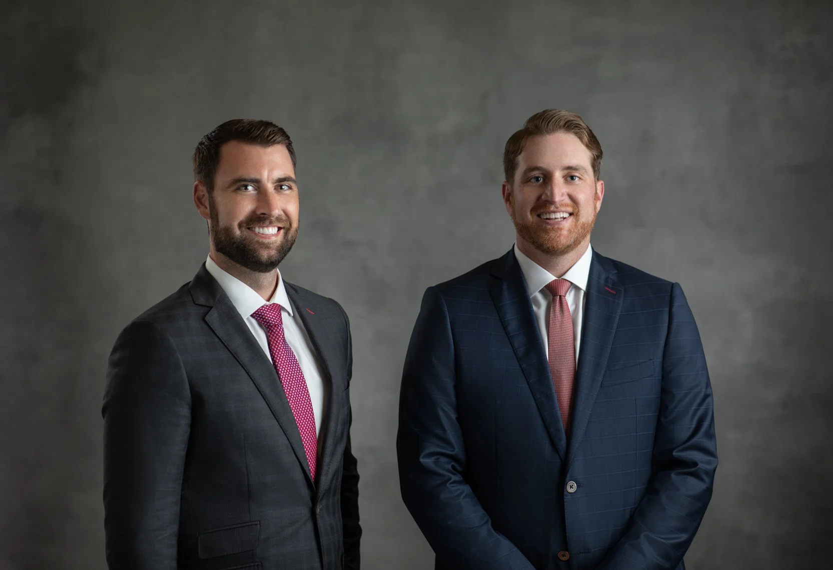 Two smiling men in suits standing in front of a plain gray background.