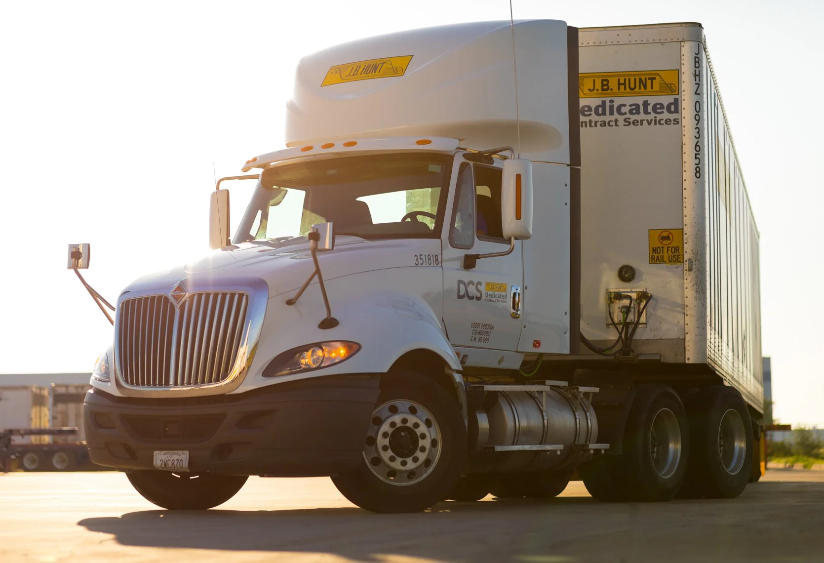 A white semi-truck with a large cargo trailer parked on a lot during sunset. The truck has the logo J.B. Hunt and text indicating it's for dedicated contract services. The truck's cab has side mirrors and a front grille, with sunlight reflecting off 