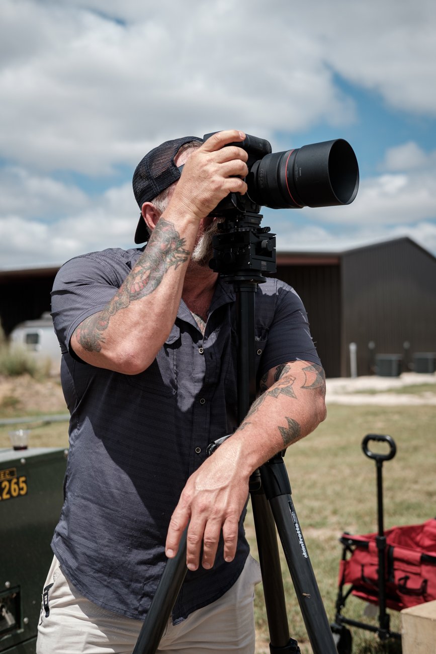 A man with tattoos wearing a baseball cap takes a photograph with a large professional camera mounted on a tripod outdoors on a cloudy day.