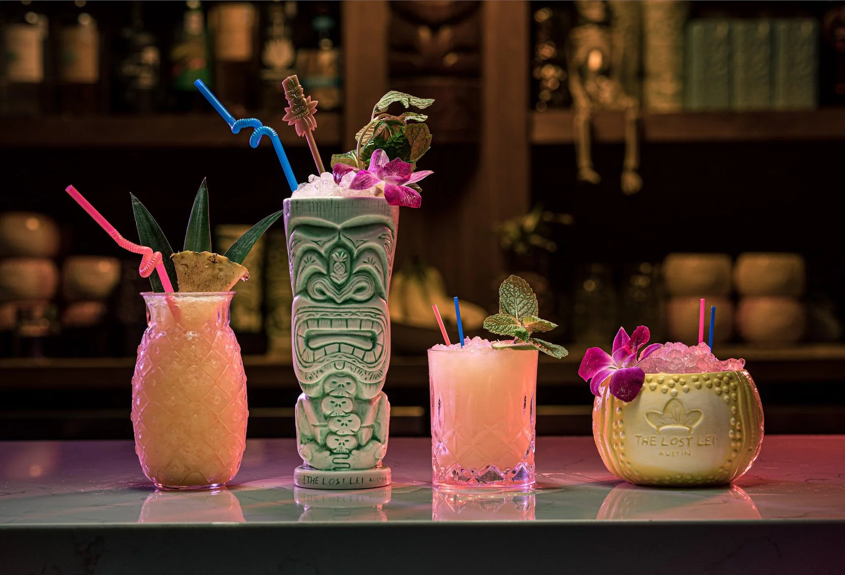 Three colorful tropical cocktails with garnishes like pineapple, mint, and flowers on a bar counter.