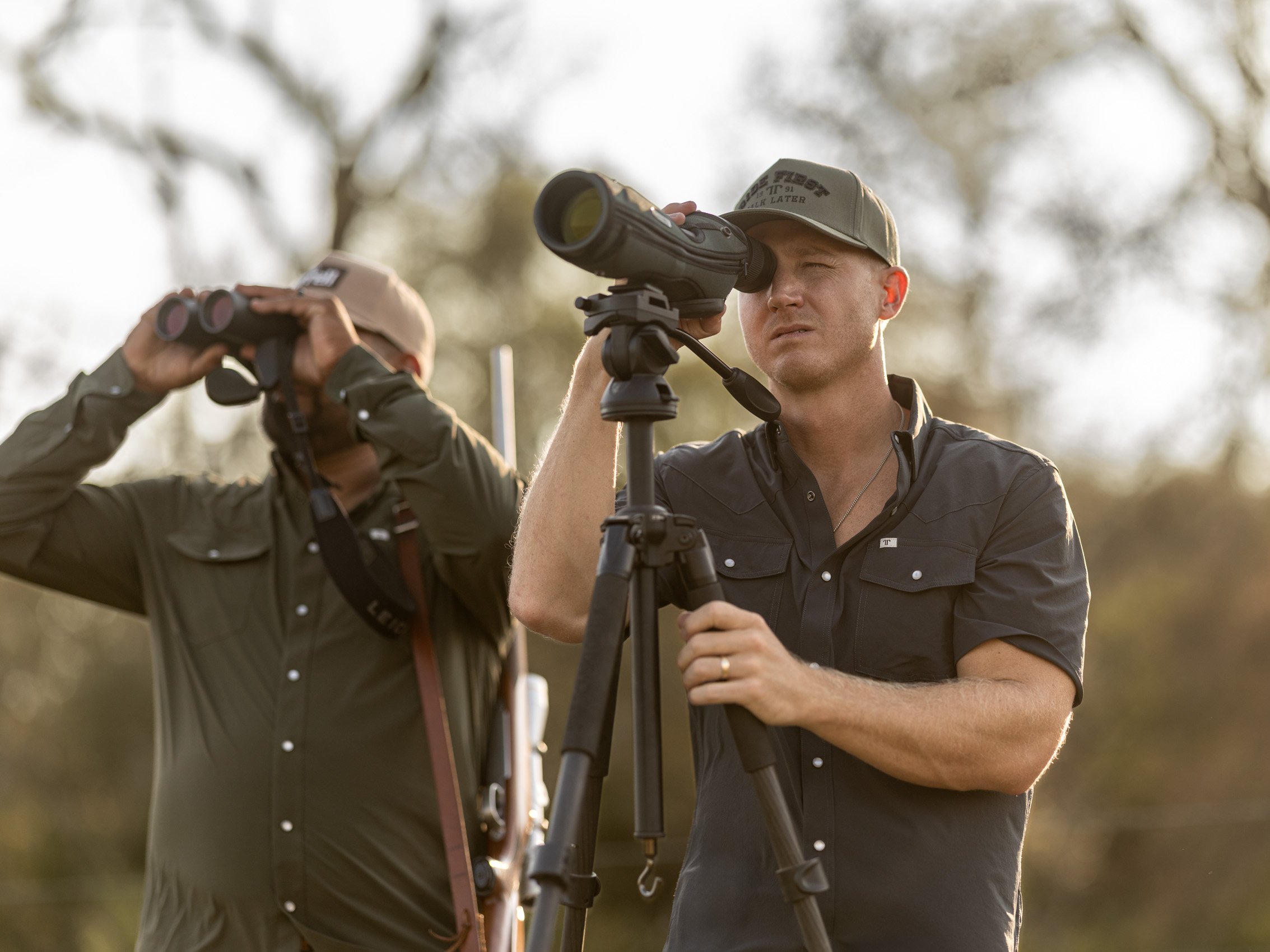 Two men outdoors, one looking through a spotting scope attached to a tripod, and the other adjusting binoculars. Both are wearing casual outdoor clothing and hats, with blurred trees in the background.