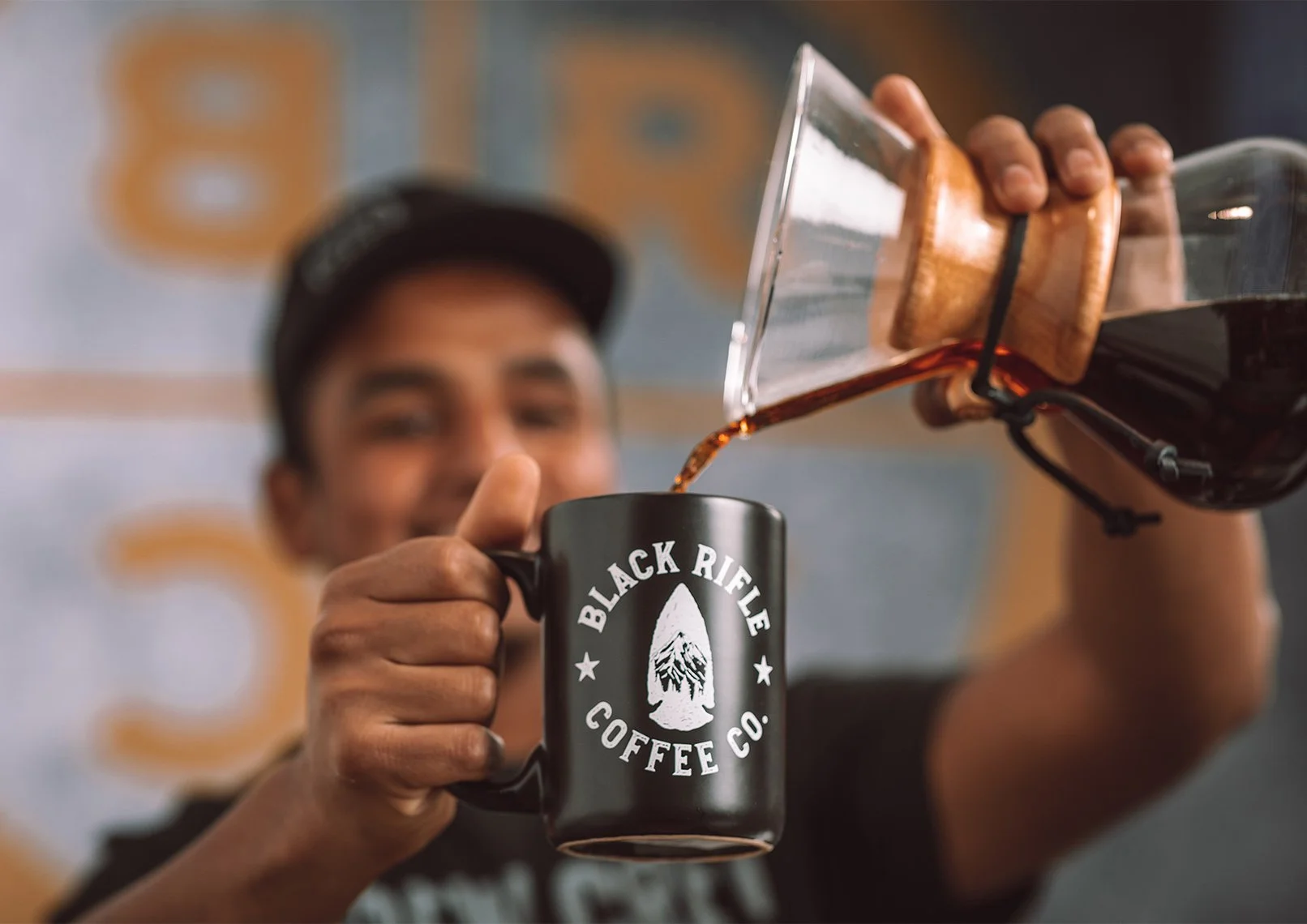A person is pouring coffee from a french press into a black coffee mug that has the logo of Black Rifle Coffee Co on it. The person is smiling and wearing a cap.