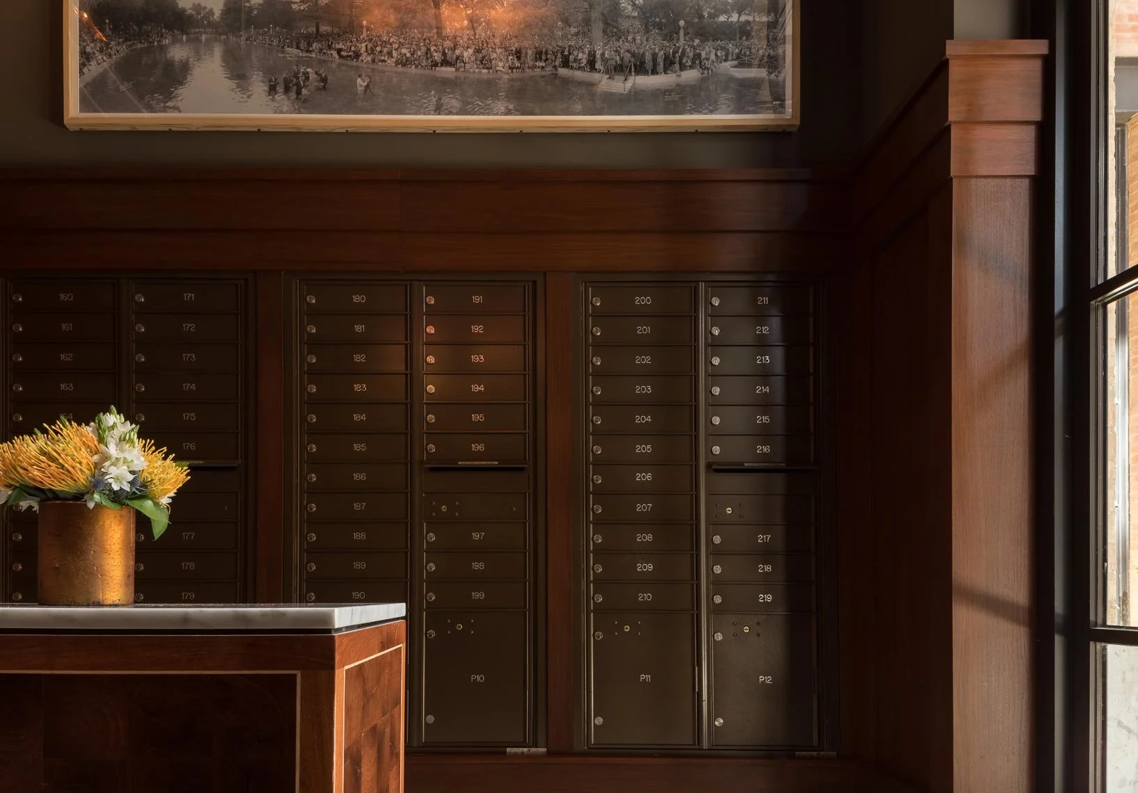 A set of black mailboxes with numbered labels inside a brick and wood interior, next to a window with sunlight. A flower arrangement in a gold vase is on a counter in the foreground, and there is a large black-and-white photograph hanging above the m