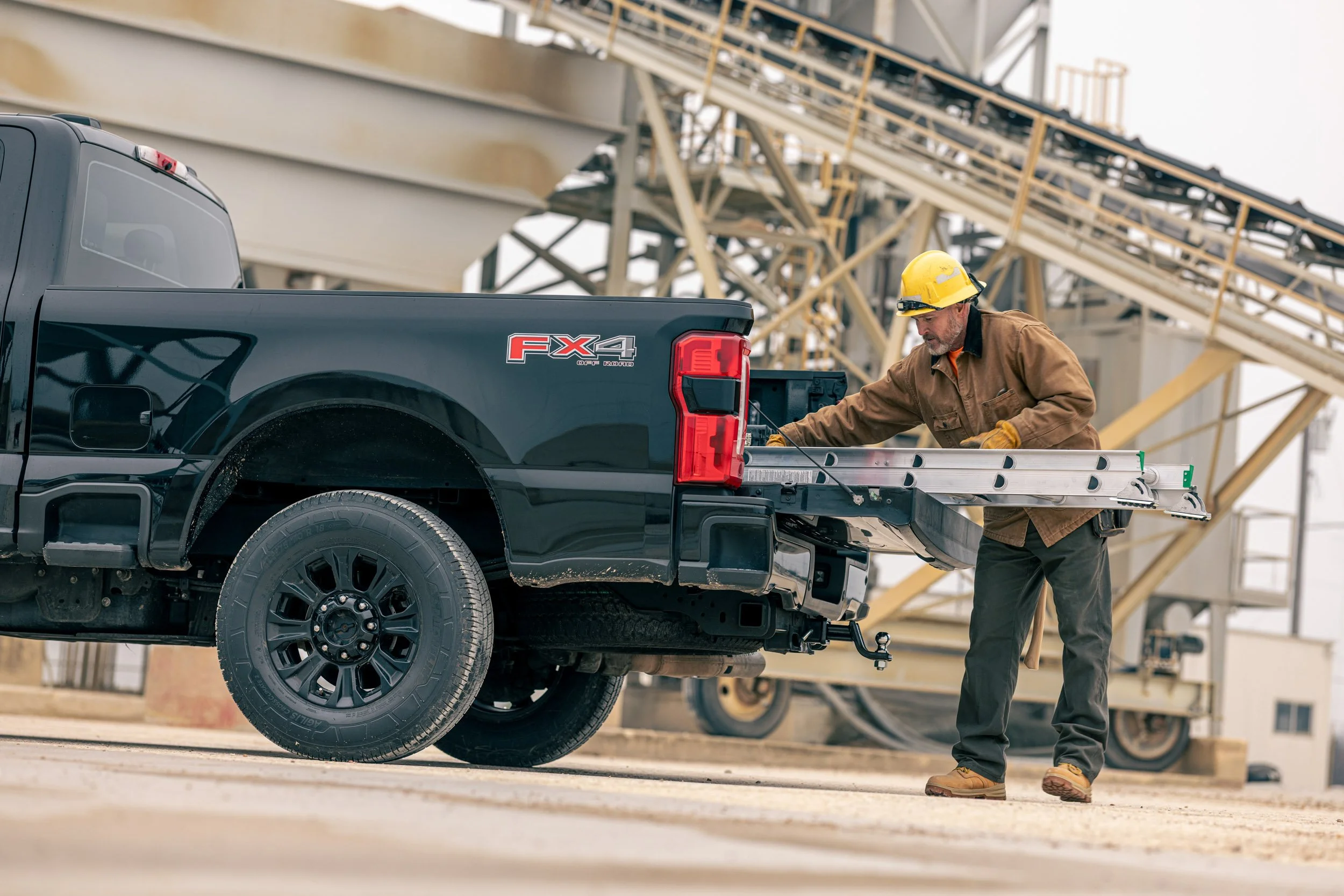 A construction worker wearing a yellow safety helmet and brown jacket loading something into the back of a black pickup truck at a construction site with metal scaffolding and structures in the background.
