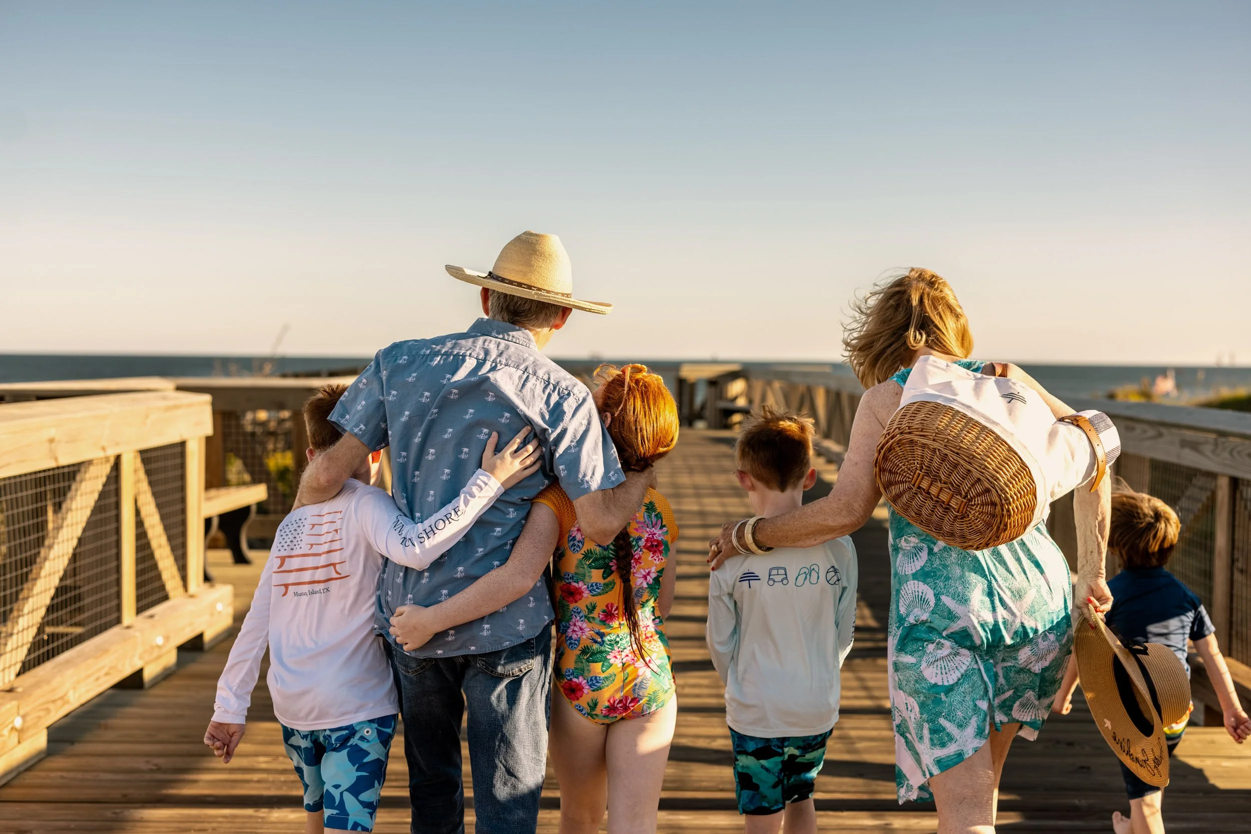 A group of people, including children and adults, gather on a wooden pier near the ocean during sunset. Many are wearing summer clothes and hats, and they seem to be enjoying a casual, outdoor social gathering in a coastal setting.