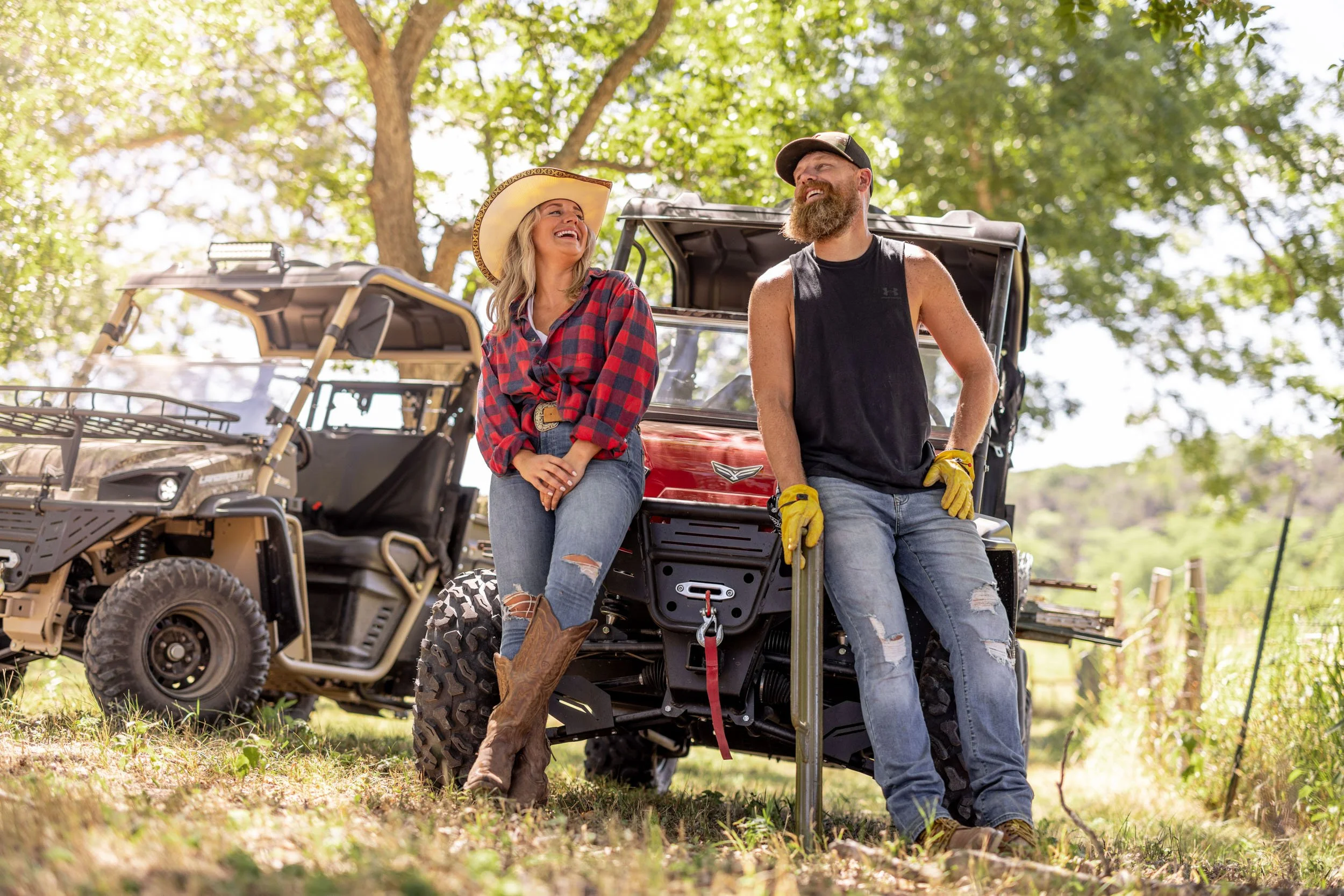 A man and woman smiling and talking outdoors, next to all-terrain vehicles, under trees in a natural setting.