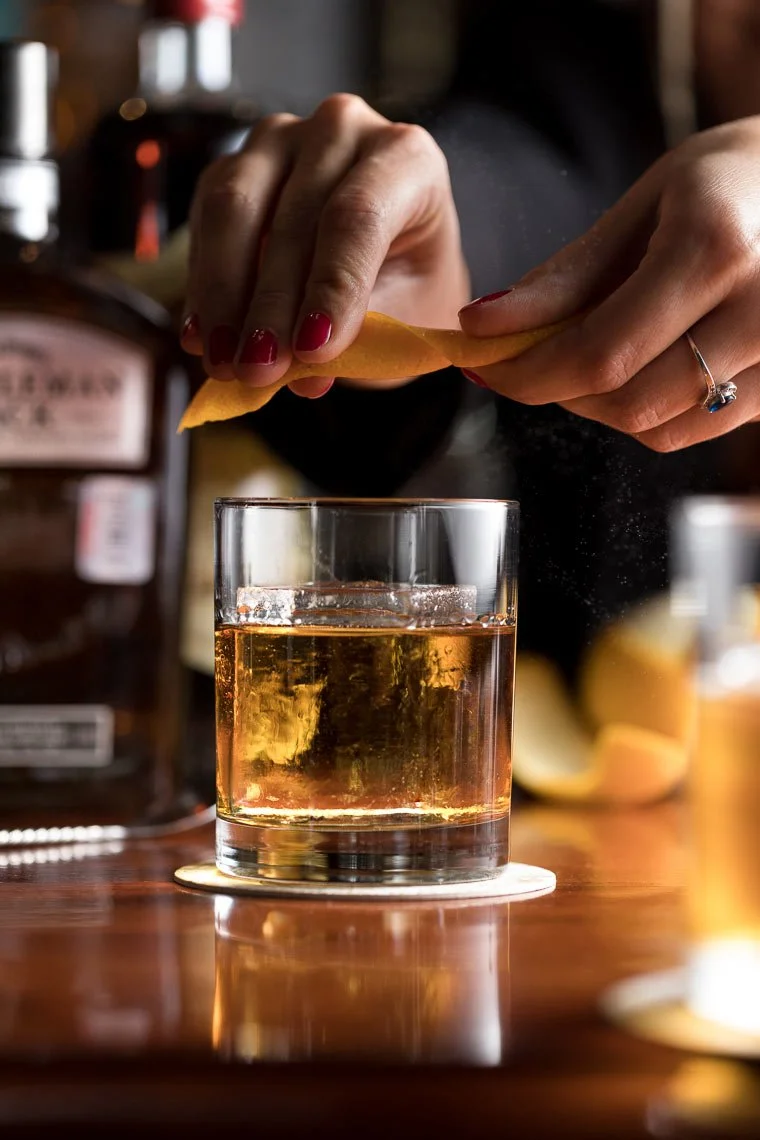 Person squeezing lemon peel into a glass of whiskey on a wooden bar counter.