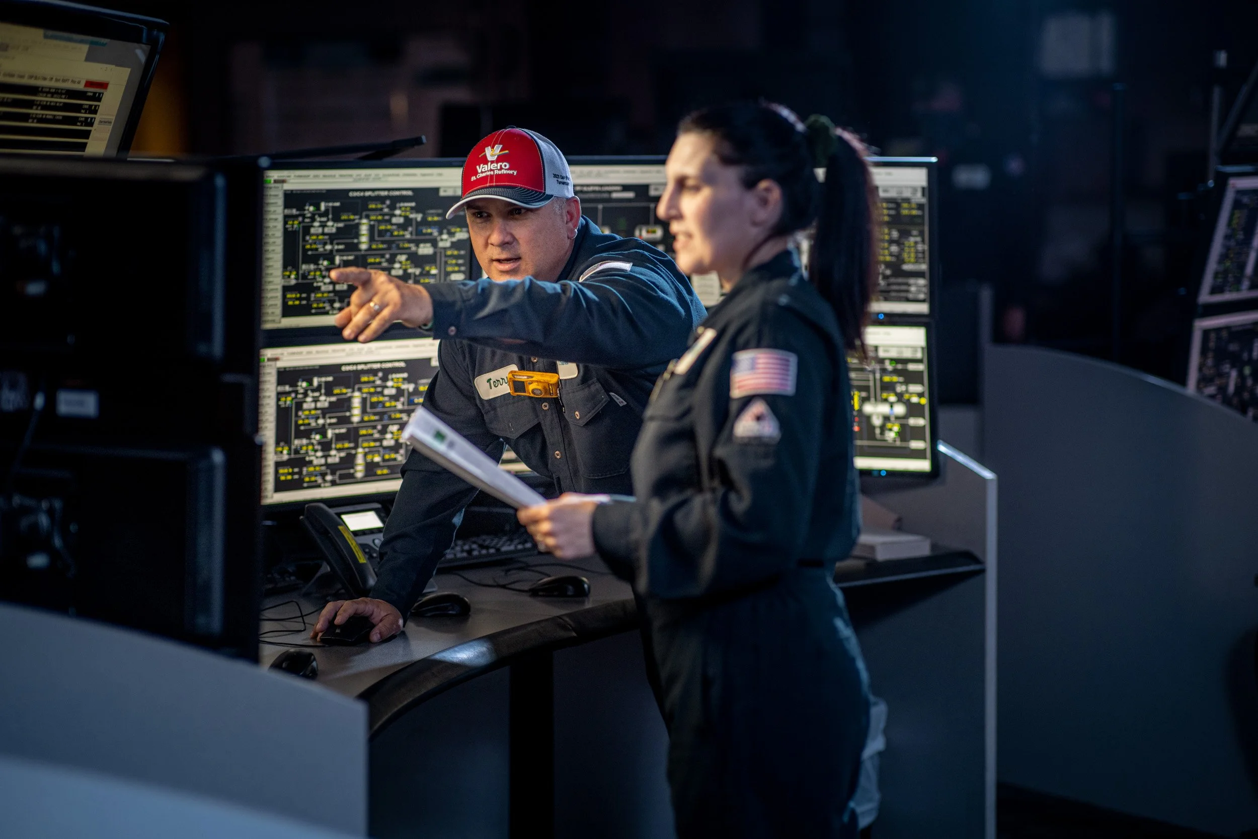 Two people, a man and a woman, in a control room or command center with multiple monitors displaying technical data. The man, wearing a red cap and pointing at one of the screens, appears to be explaining something to the woman, who is holding a note