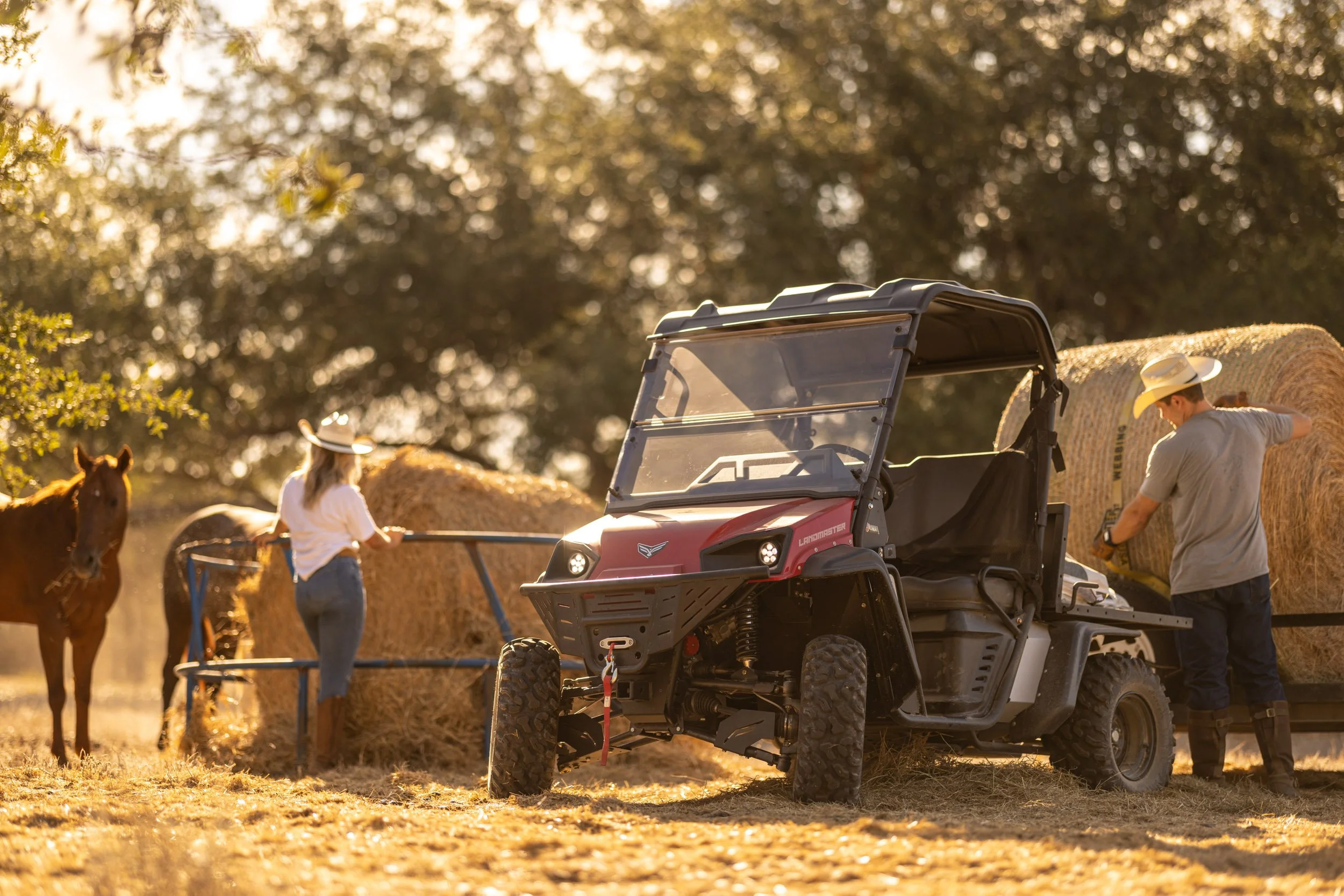 People working with hay bales using an off-road utility vehicle in a rural setting during sunset.