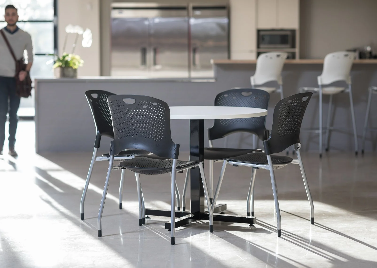 Empty round table with four black plastic chairs in a bright, modern kitchen or cafe setting.