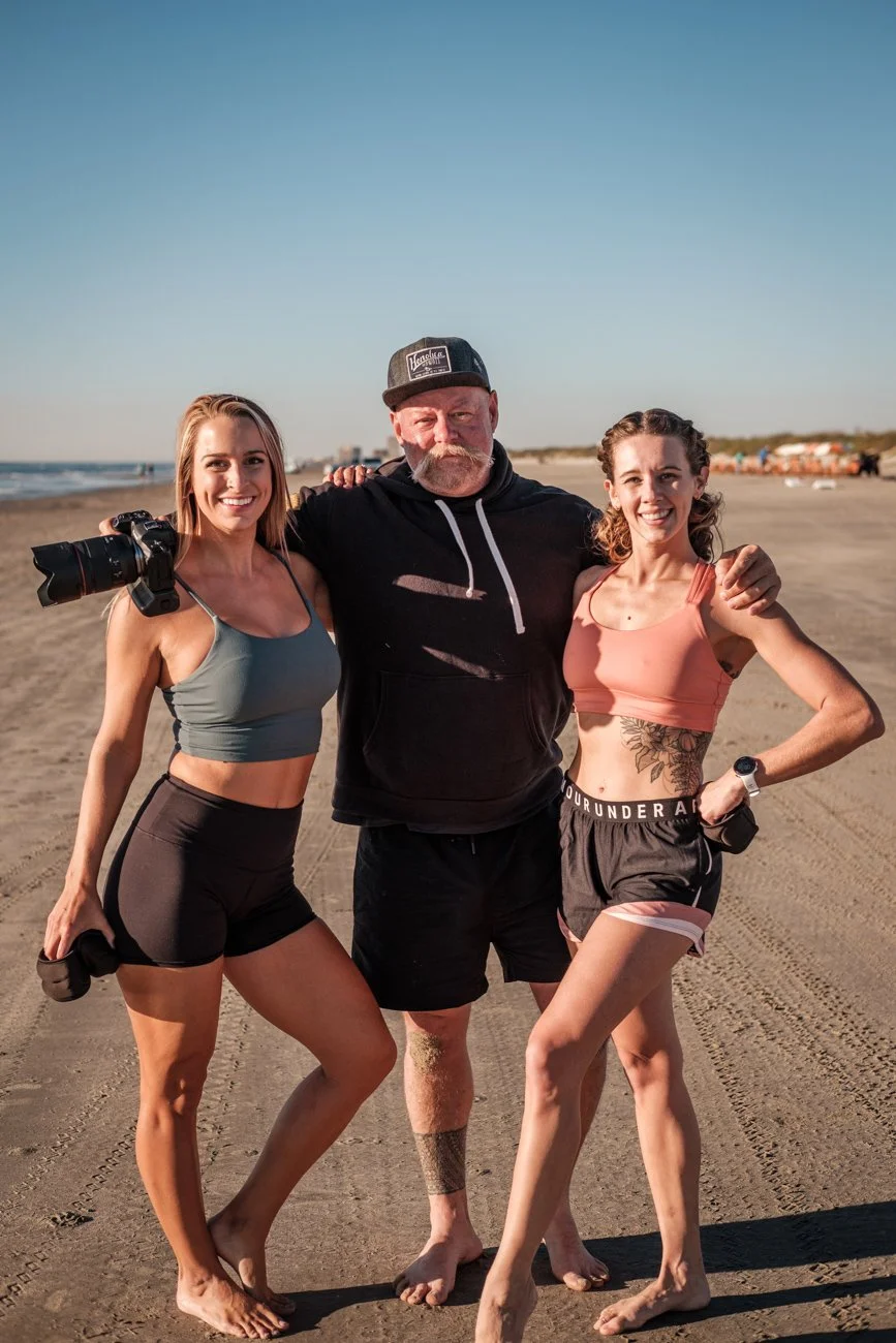 Three people standing on a beach, smiling at the camera, with the ocean and sky in the background. The woman on the left is holding a camera, wearing a sports bra and shorts. The man in the middle wears a black hoodie, shorts, and a cap. The woman on