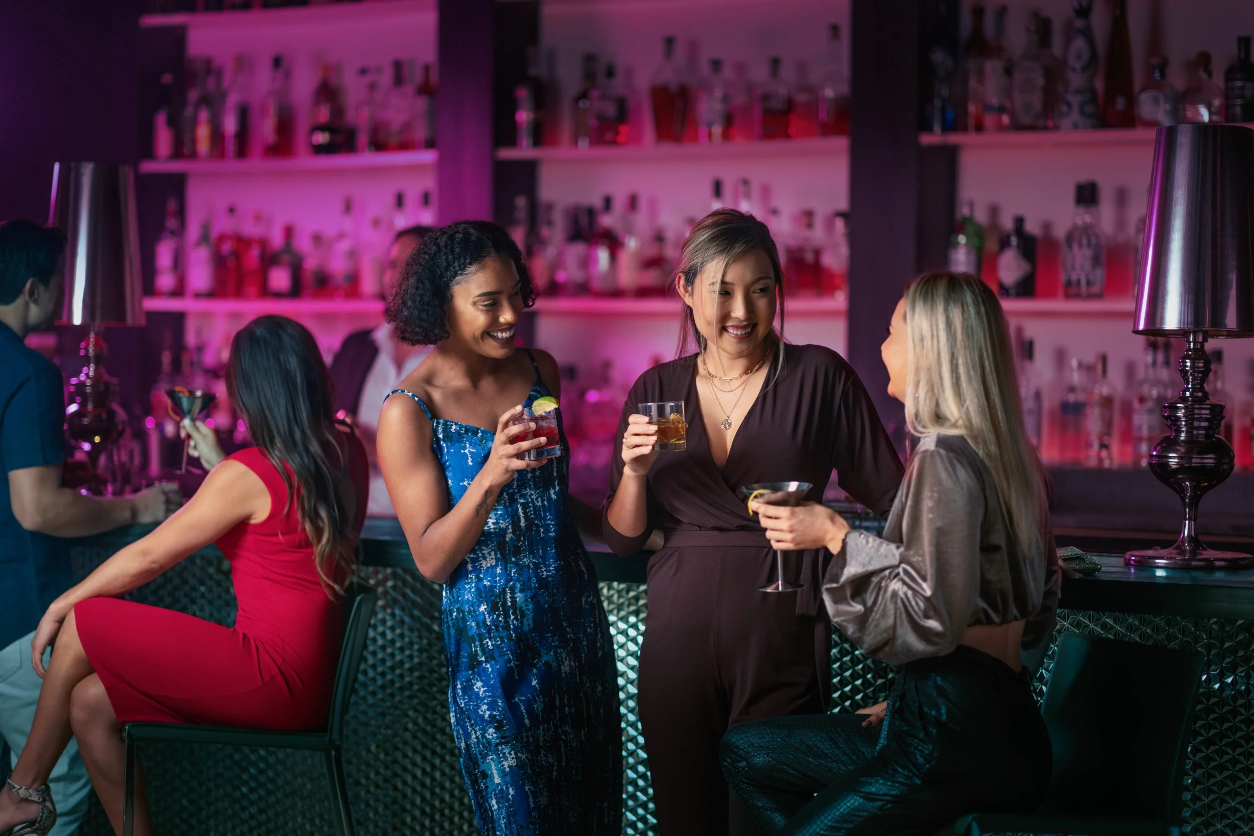 A group of women socializing and holding drinks at a bar with pink and purple lighting.