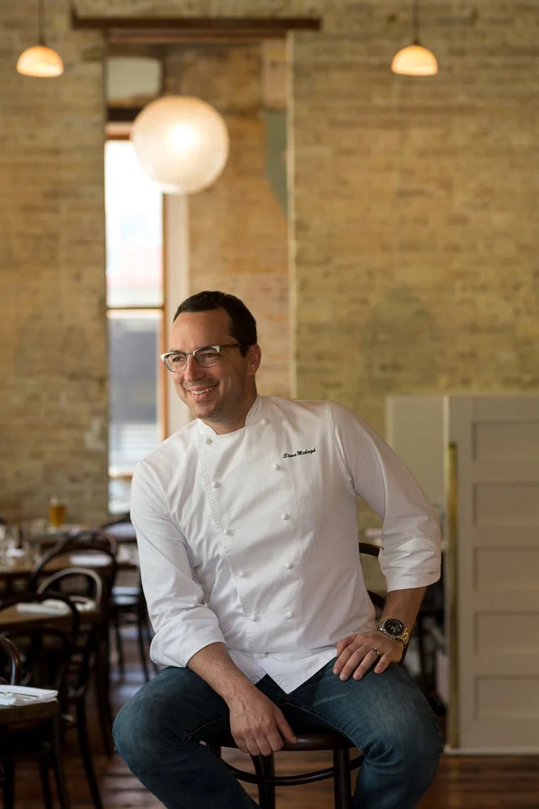 A man in a white chef's coat sitting on a stool in a restaurant, smiling, with brick walls and hanging lights in the background.