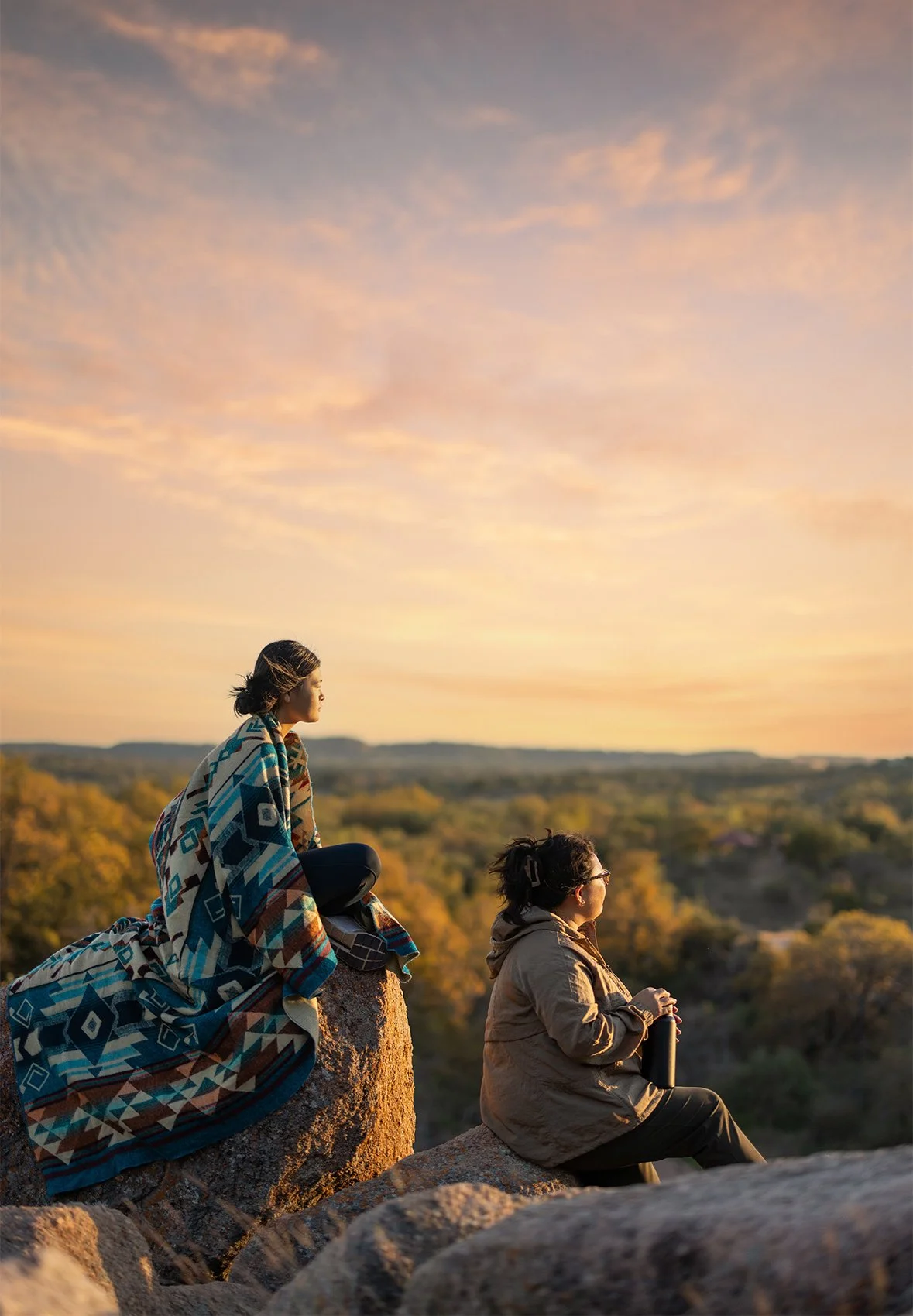 Two women sitting on rocks outdoors during sunset. One woman with dark hair, wearing a patterned blanket, is kneeling and looking into the distance. The other woman with glasses and dark hair tied back, wearing a brown jacket, is sitting with a therm