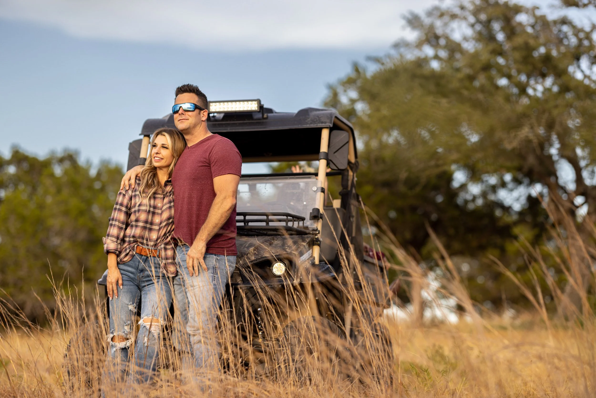 A man and woman standing together outdoors in a field with tall grass, near a side-by-side off-road vehicle, with trees and a partly cloudy sky in the background.