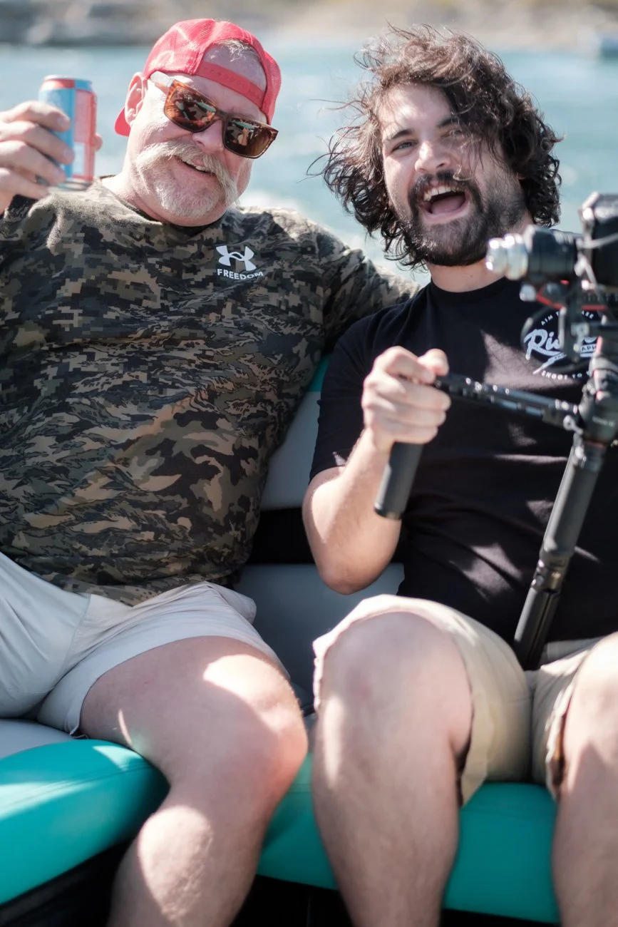 Two men enjoying a boat ride, one with sunglasses and a red cap, the other with curly hair and a beard, smiling and holding a camera.