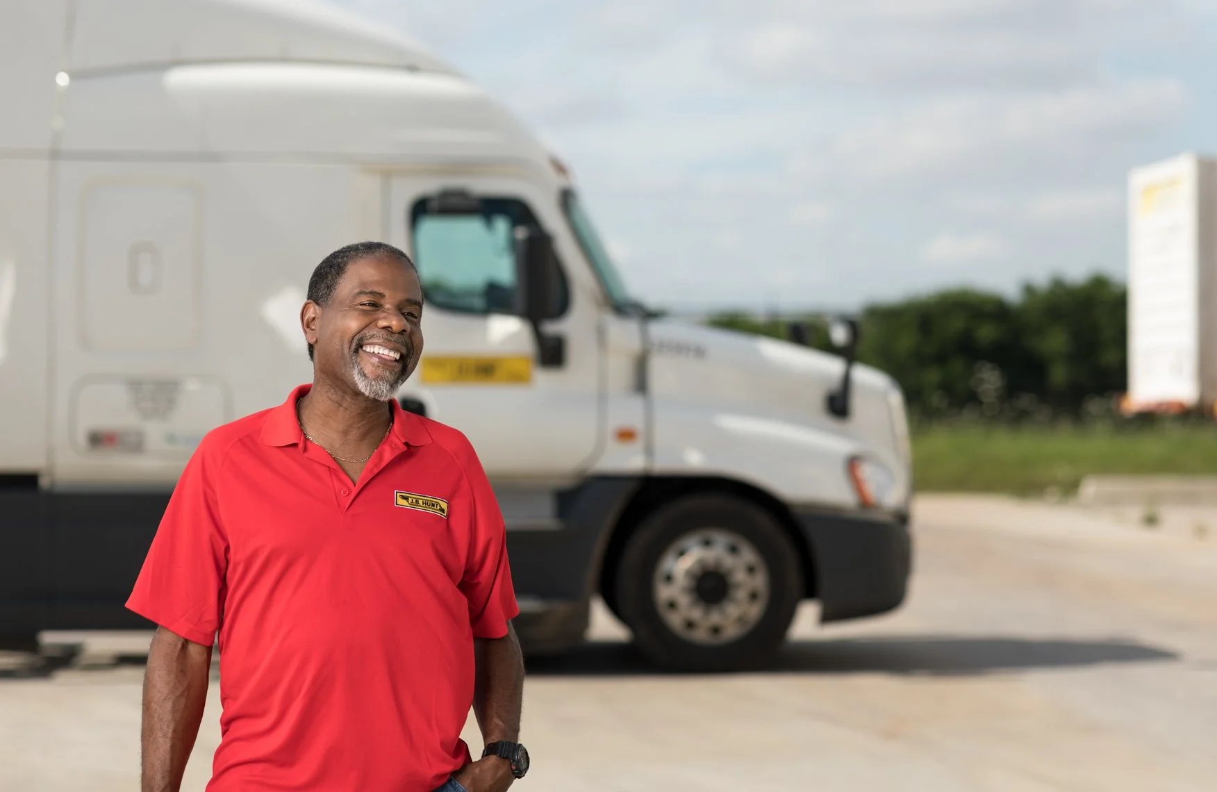 A smiling man in a red shirt standing in front of a white semi-truck in an outdoor setting.