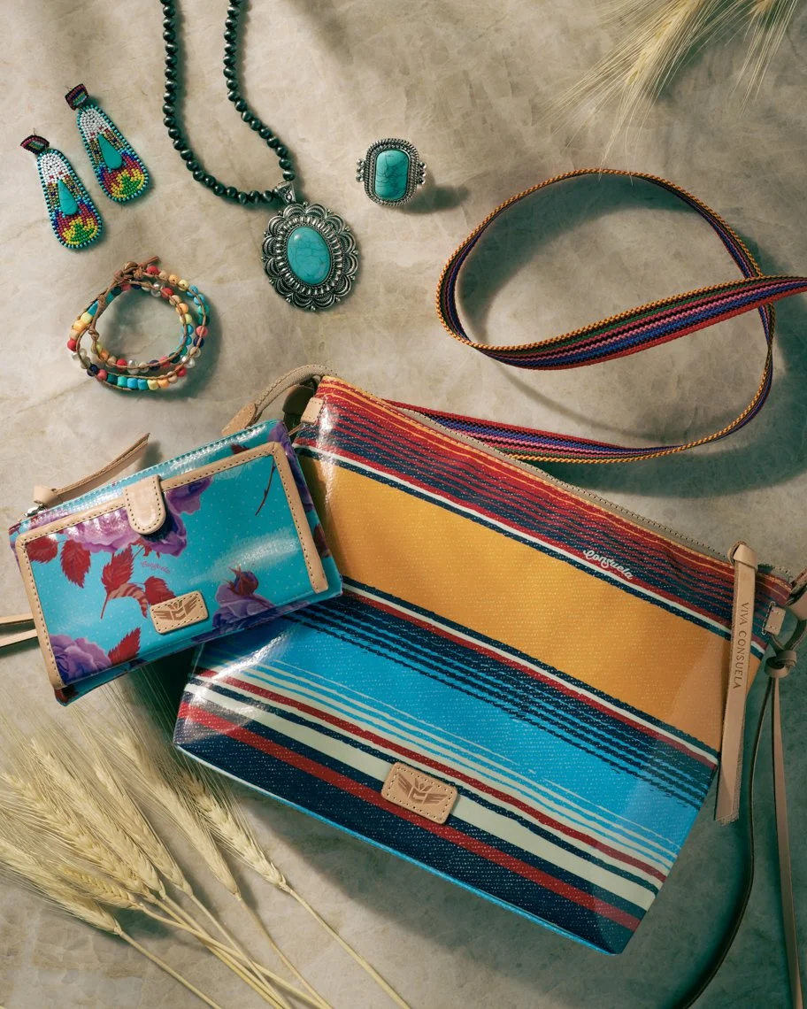 Various colorful accessories including turquoise jewelry, a multicolored striped tote bag, a matching wallet, beaded jewelry, and a striped fabric strap, arranged on a light surface with some wheat stalks and dried leaves.