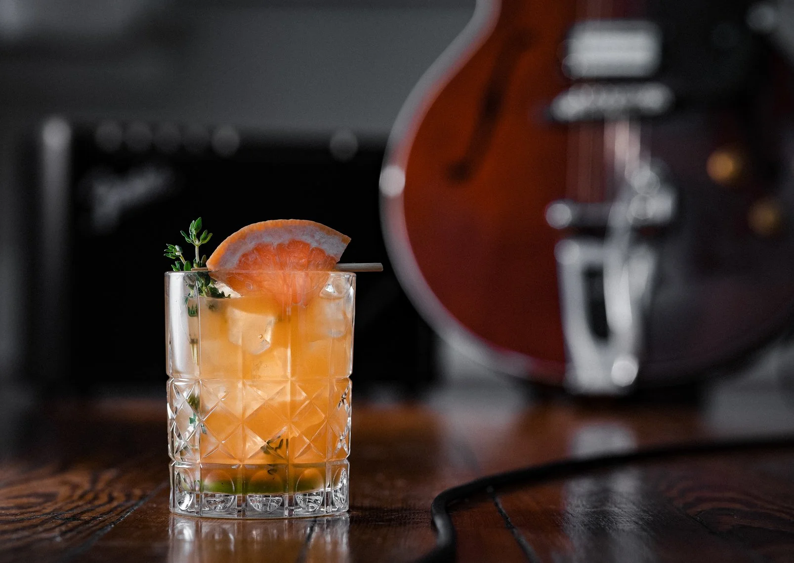 A glass of orange cocktail garnished with a slice of orange and a sprig of thyme, placed on a wooden surface, with a blurred microwave oven in the background.