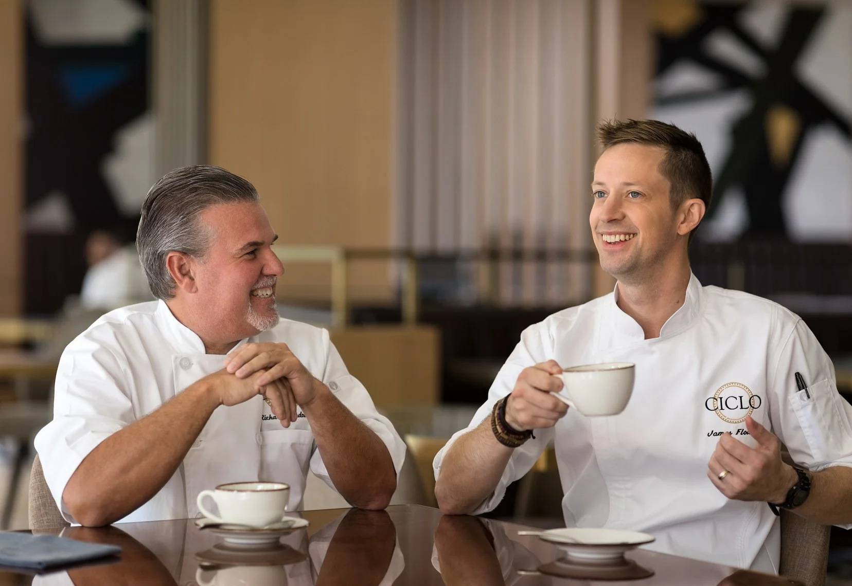 Two male chefs in white coats smiling, sitting at a table with coffee cups, having a conversation in a restaurant or cafe.