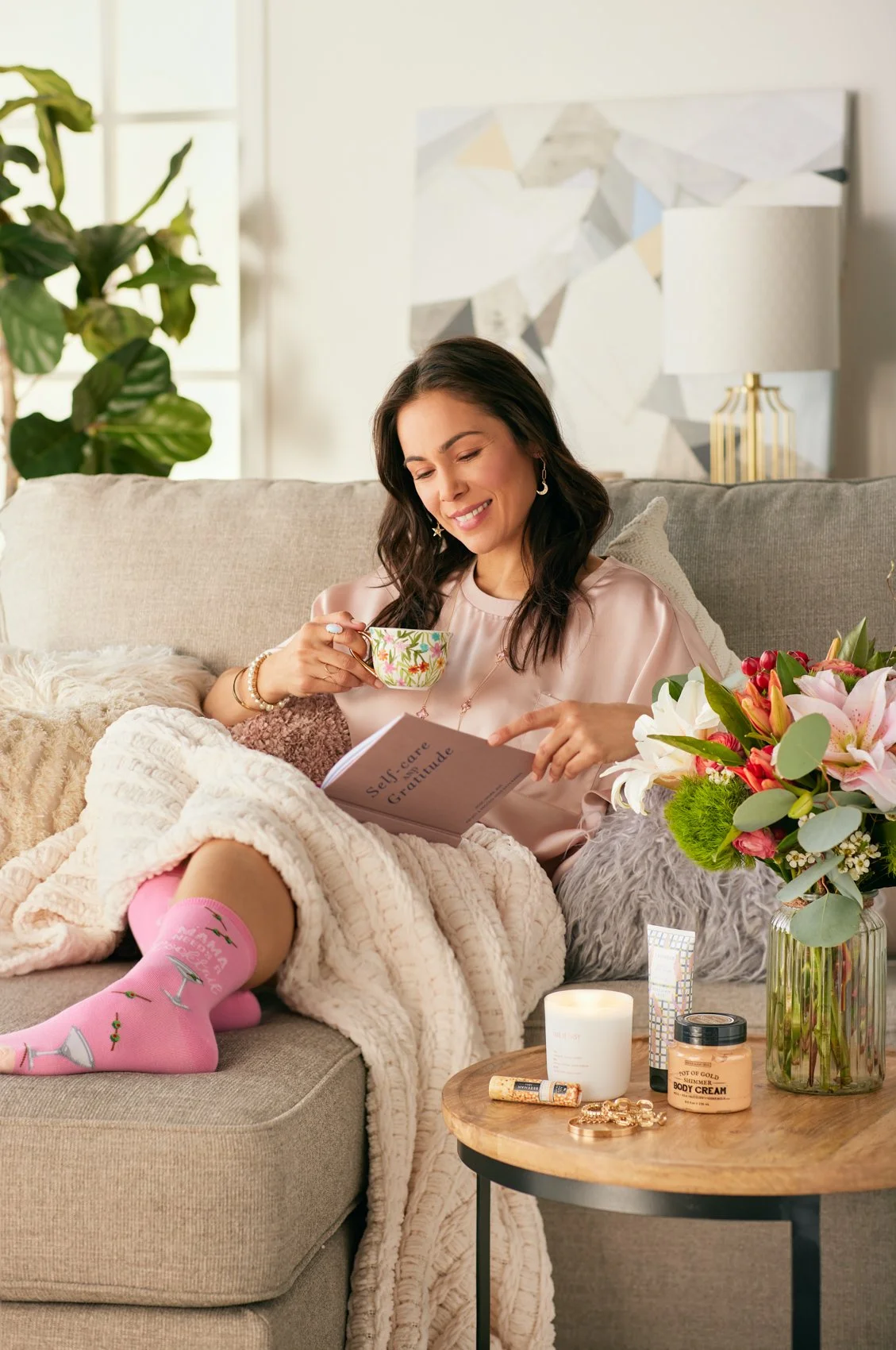 A woman sitting on a beige sofa with a book titled 'Self-care Gratitude,' holding a floral teacup, with a blanket, flowers, and skincare products nearby.