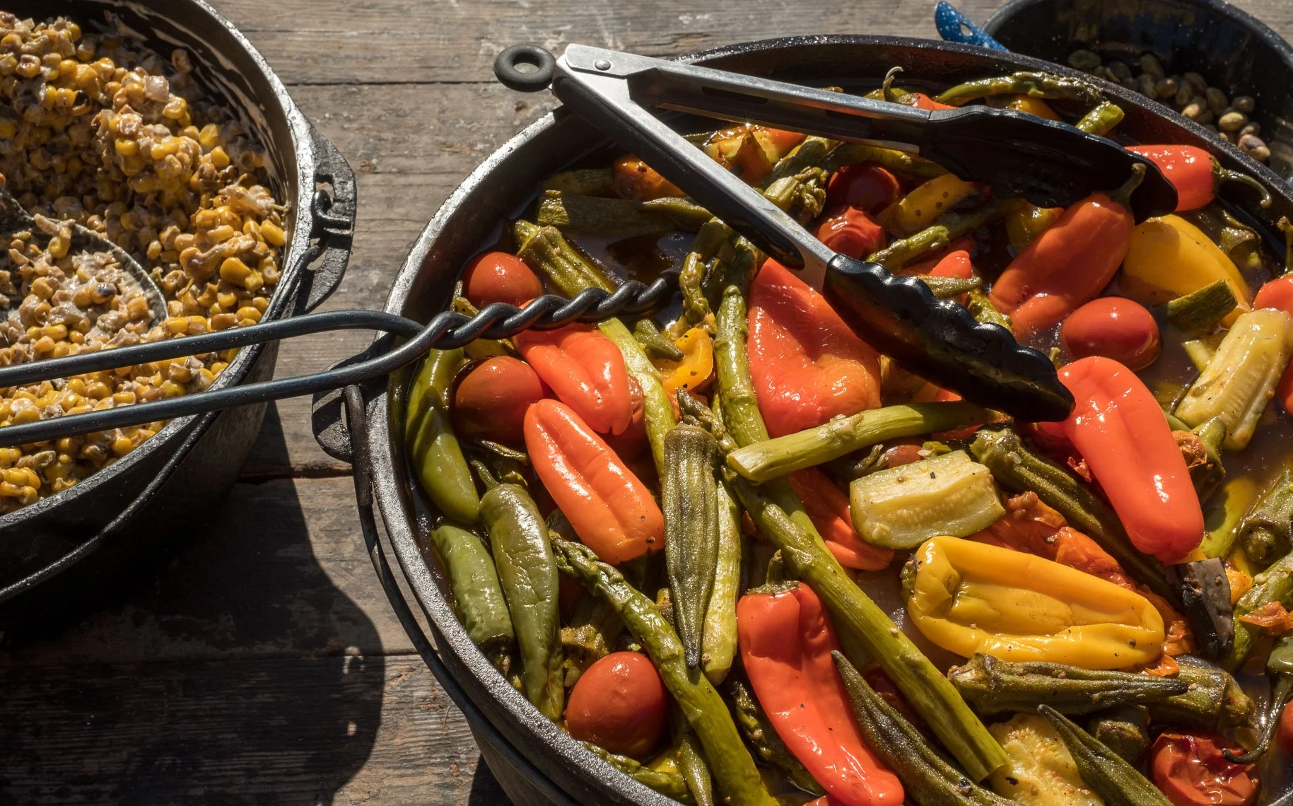 Cast iron skillet filled with cooked mixed vegetables including green beans, cherry tomatoes, yellow and orange peppers, and zucchini. Tongs rest on top of the vegetables. To the left, there is a cast iron skillet filled with cooked corn and ground m