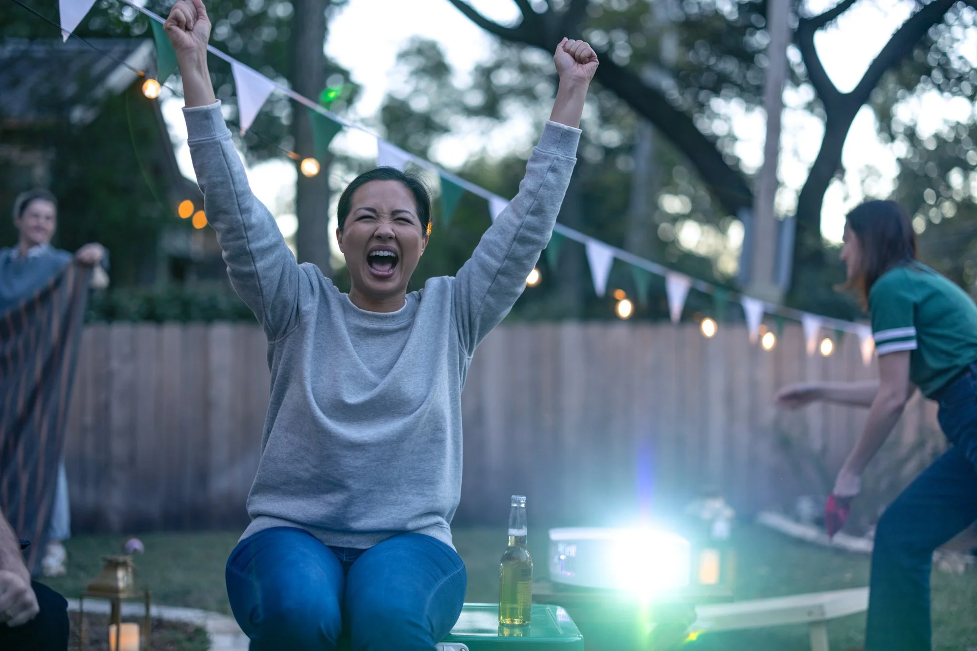 A woman with dark hair, wearing a gray sweatshirt, sitting at a table outdoors during evening or night, celebrating with her arms raised and fists clenched in excitement. There are string lights and pennant banners in the background, and a few other 