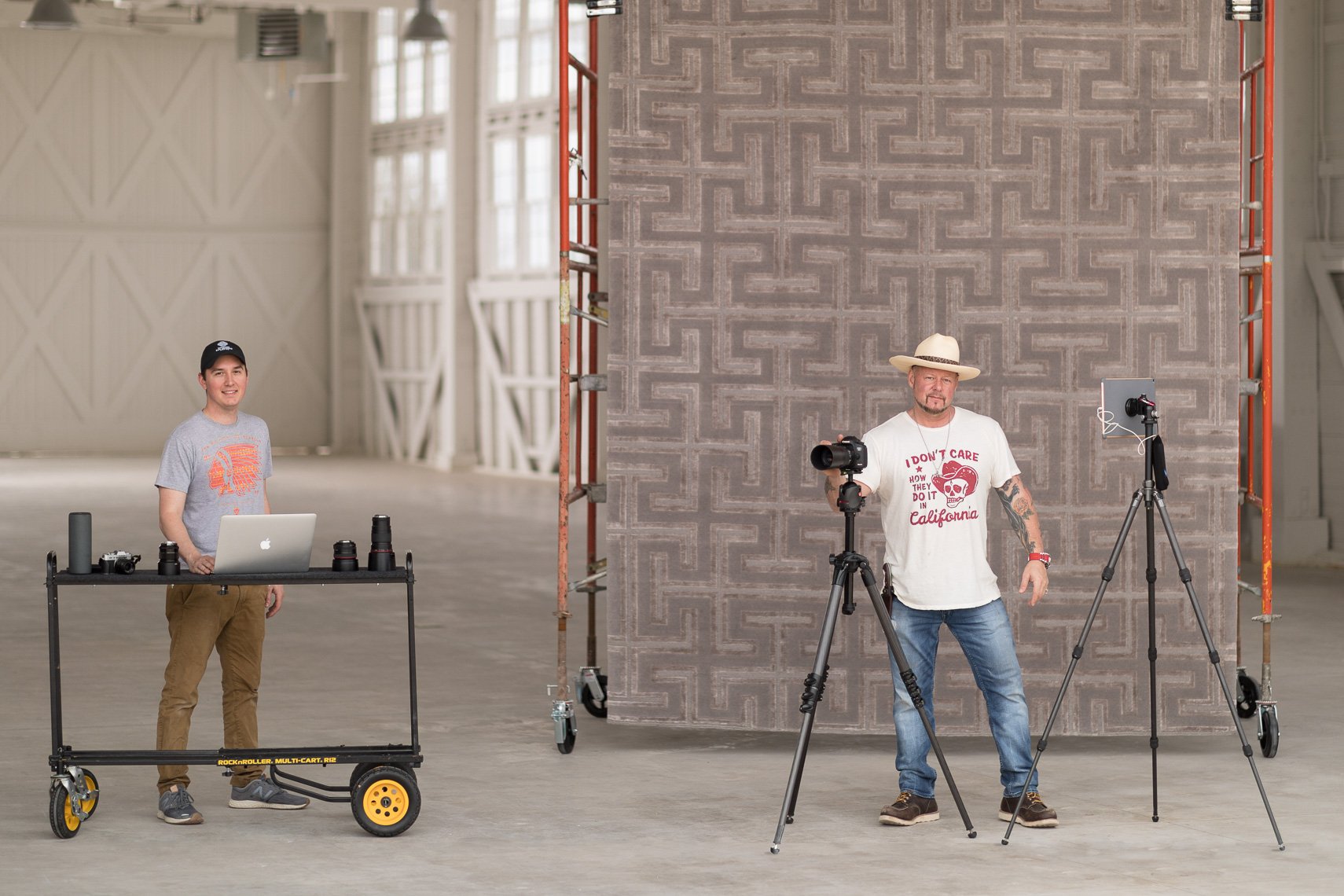 Two men in a spacious, industrial-style room with high ceilings and large windows. The man on the left is standing behind a rolling table with a laptop, camera lenses, and other equipment, wearing a gray t-shirt and tan pants. The man on the right is