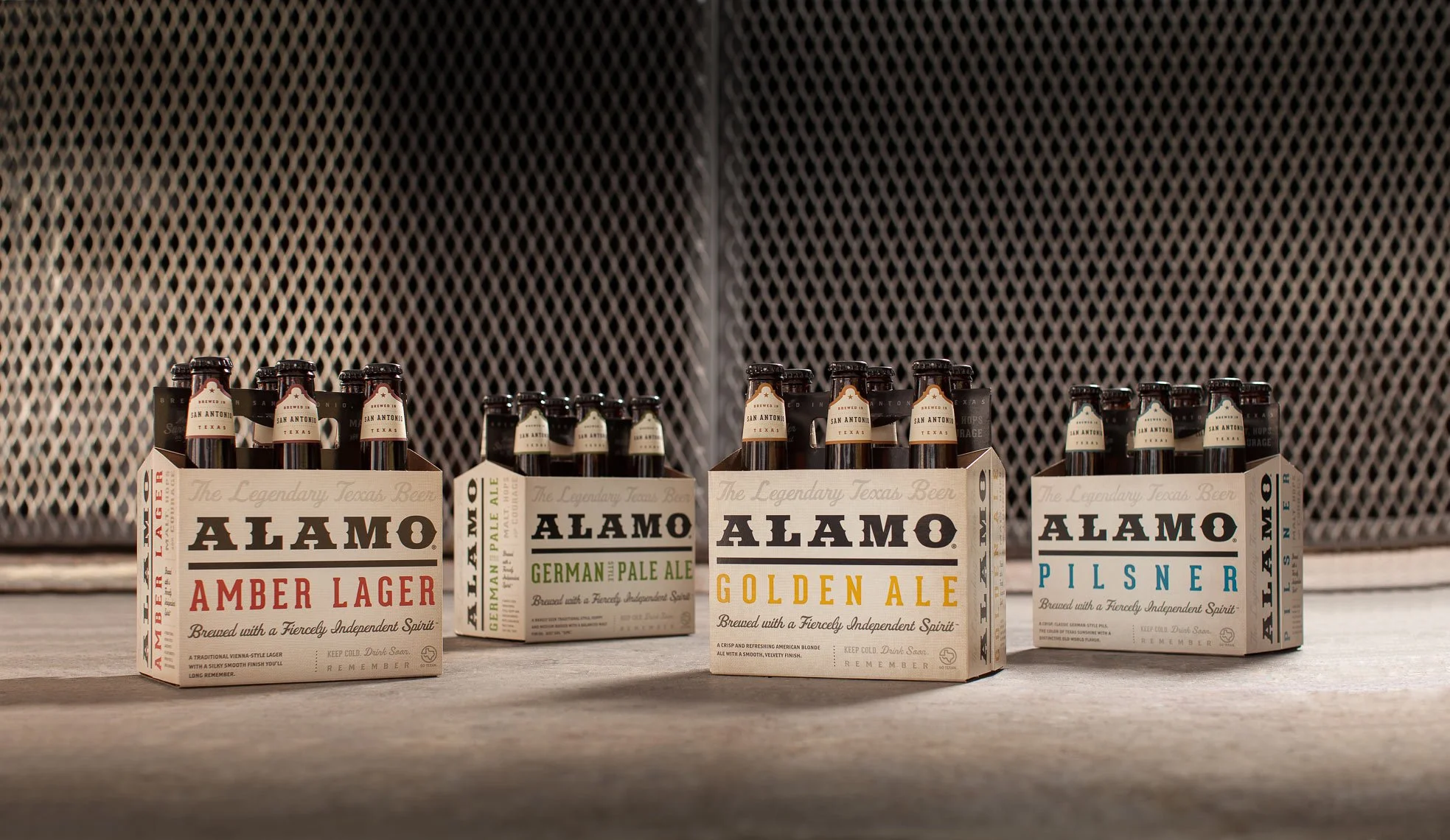 Six six-pack containers of Alamo beer, each labeled with different beer styles, arranged on a surface against a textured metallic background.