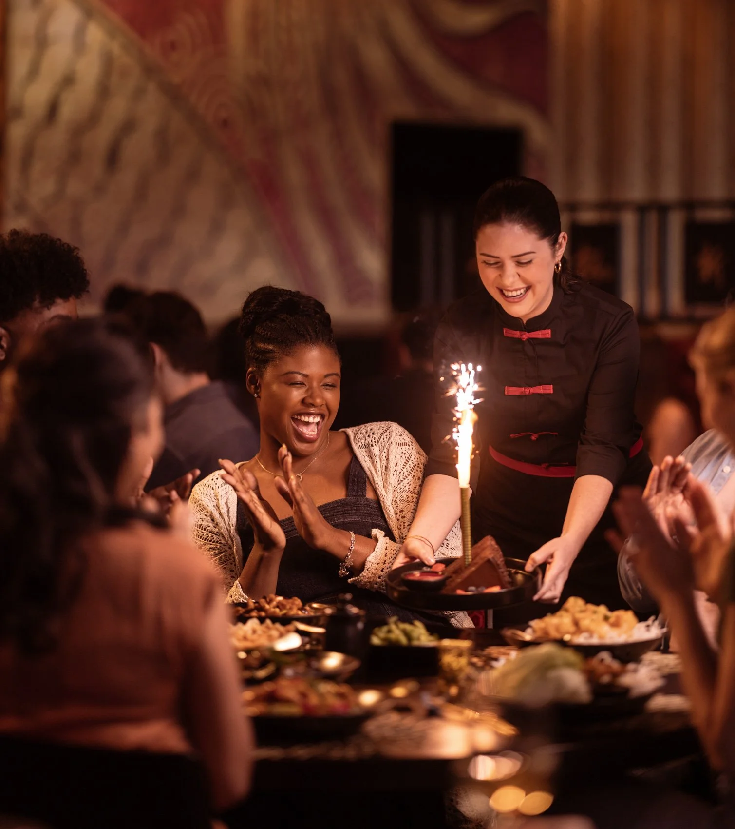 A group of people celebrating around a table at a party with a woman bringing a cake with a sparkler on top, while others smile and cheer.