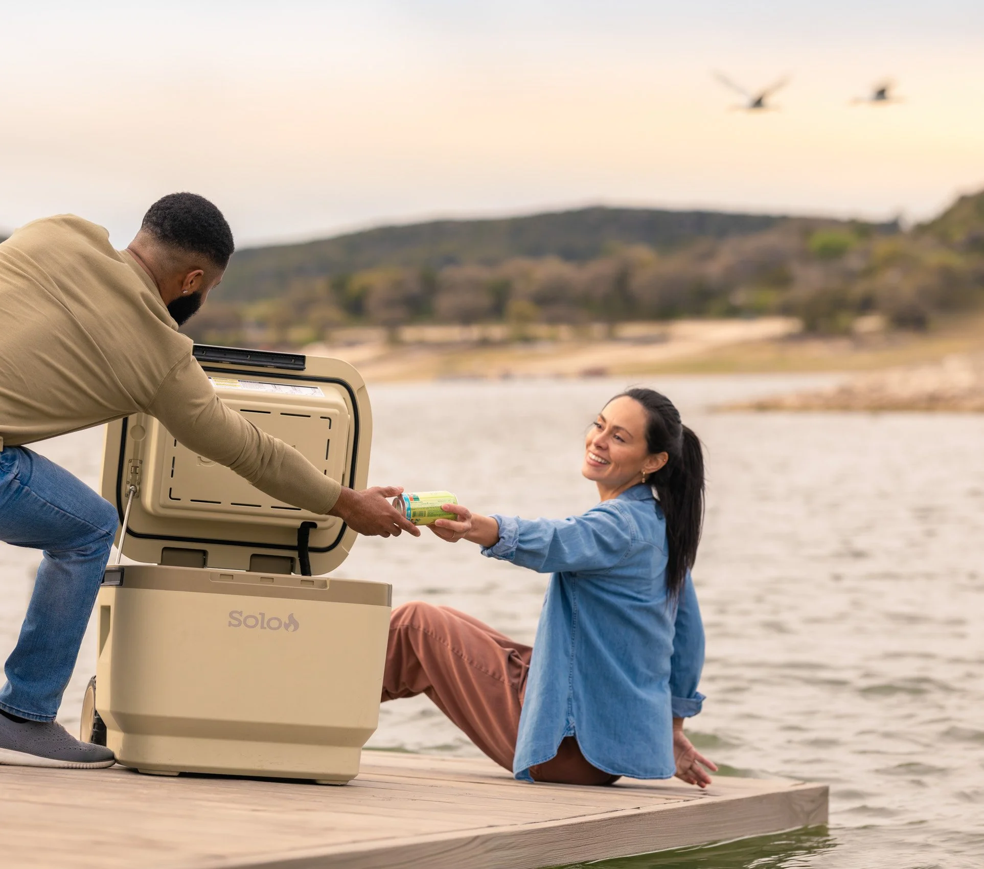 A woman with long dark hair in a ponytail, smiling, is being handed a can by a man. They are near a body of water with hills in the background, and a dog is laying on a wooden dock. The scene is outdoors, during daytime.