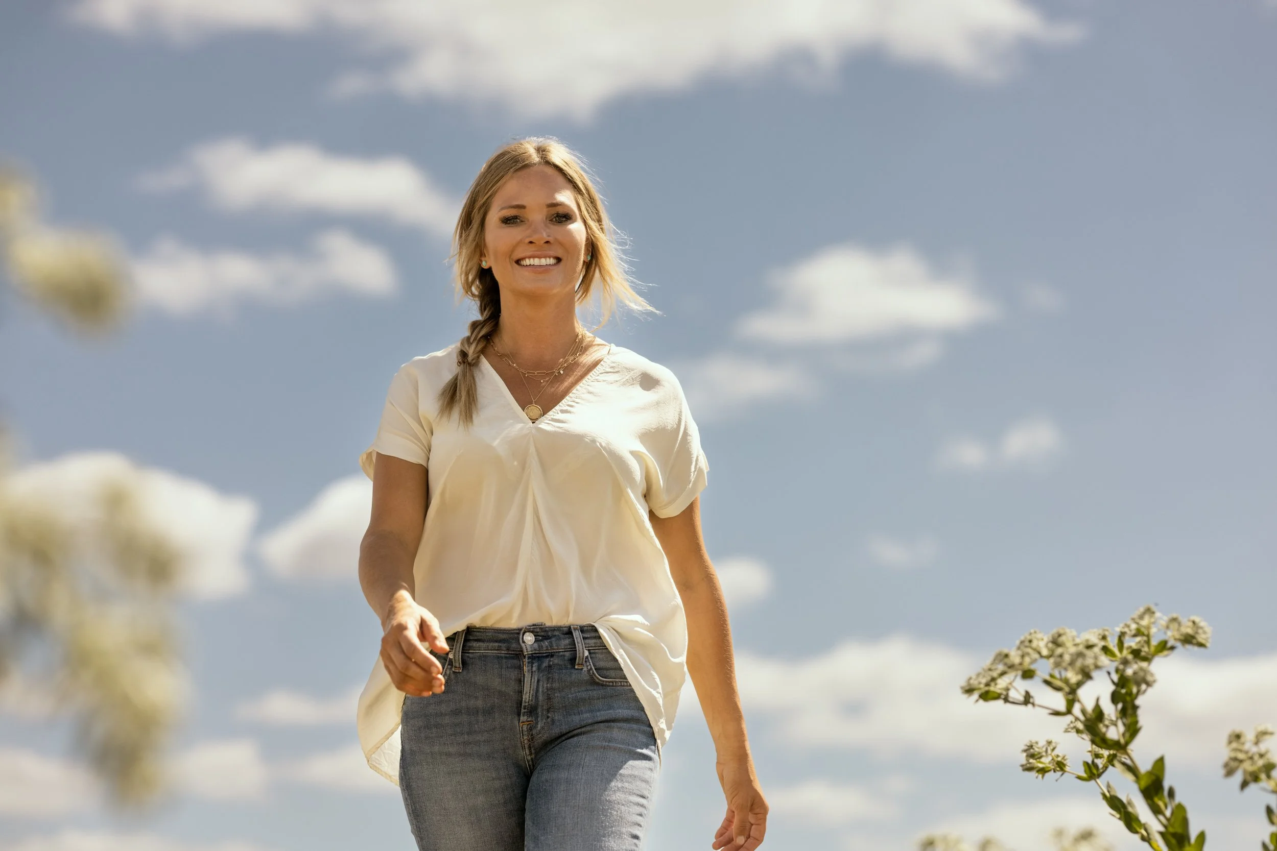 A woman with blond hair in a braid, wearing a cream-colored top and jeans, walking outdoors on a sunny day with blue skies and scattered clouds.