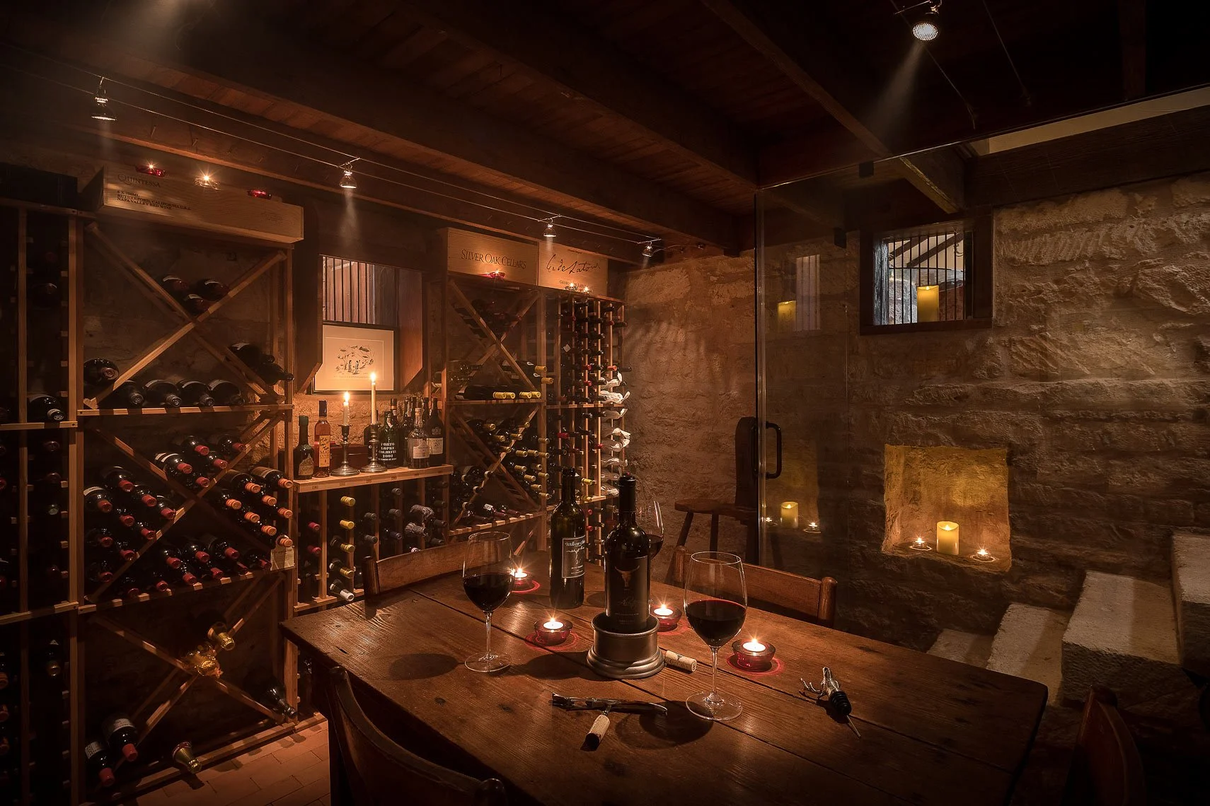 A cozy wine cellar with wooden wine racks filled with bottles, a wooden table set with two glasses of red wine and candles, candles placed along a stone shelf, and a small window with bars in a stone wall.
