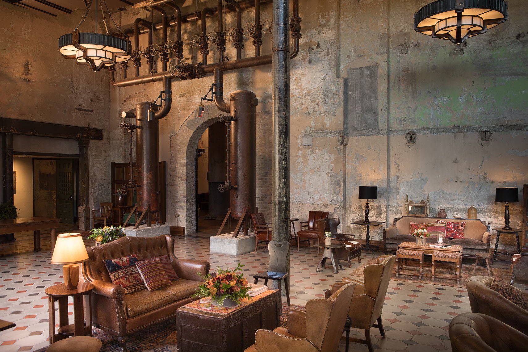 A vintage-style lounge area with leather and fabric armchairs, wooden side tables, and floral decorations, set against an industrial wall with exposed pipes and valves, and lit by large art deco ceiling lights and table lamps.