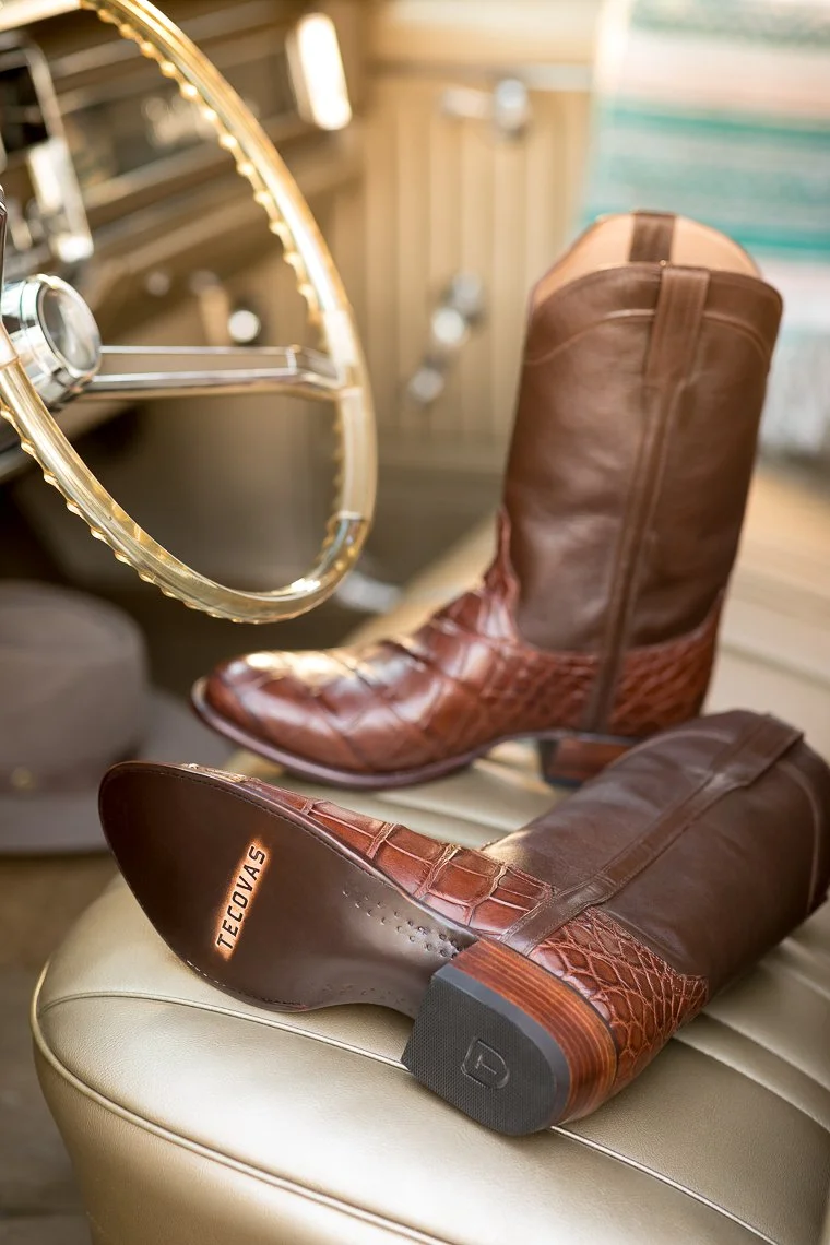 Pair of brown leather cowboy boots with crocodile texture, placed on a beige leather surface near a vintage steering wheel in the background.