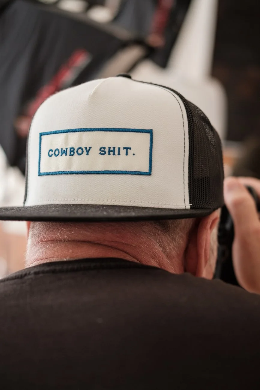 Back of a person's head wearing a trucker hat with a white front panel and black mesh side. The hat has a rectangular patch with blue stitching that reads 'COWBOY SHIT.'