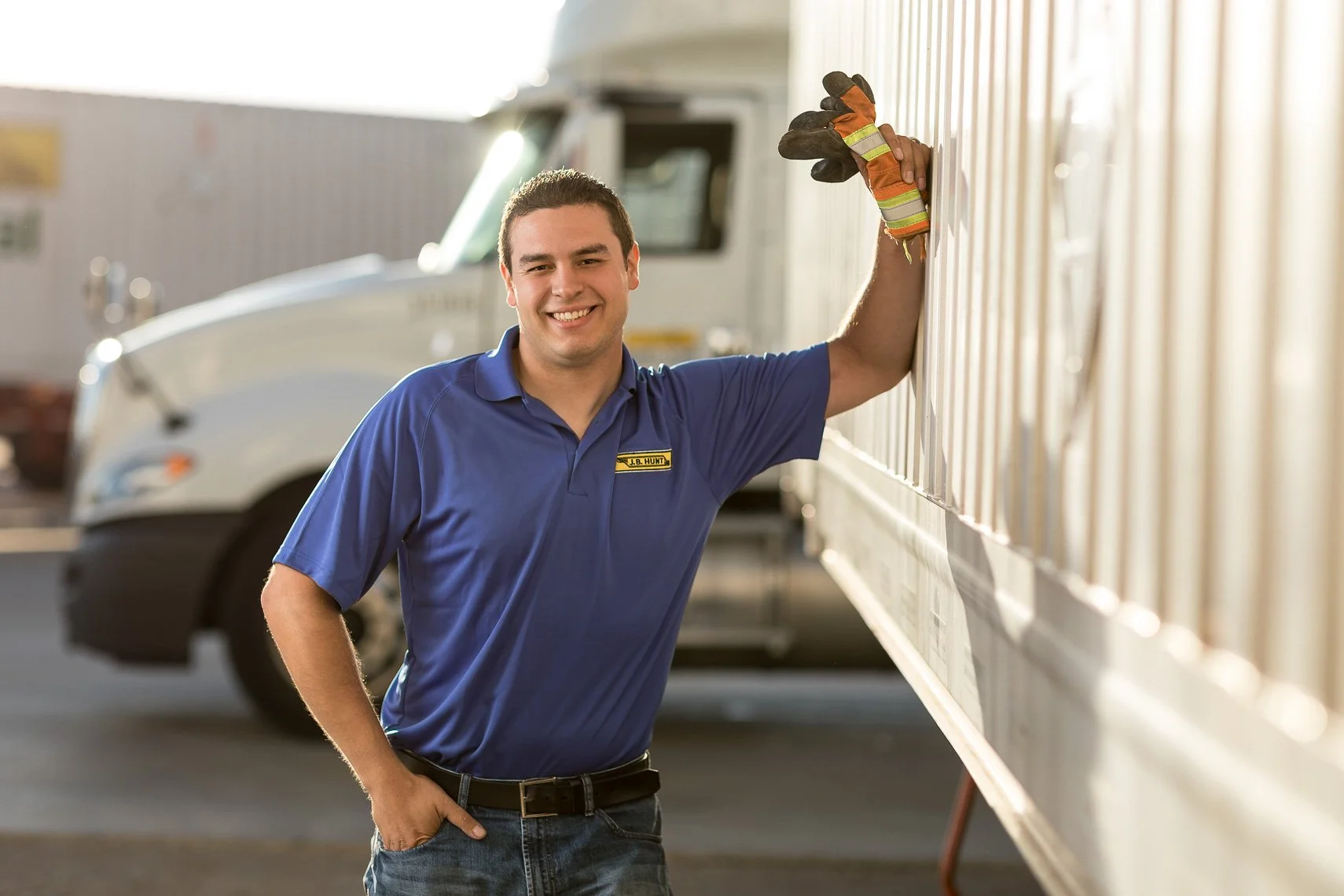 A man in a blue uniform with a name tag that says "J.B. Hunt" is smiling and standing next to a large truck trailer, leaning on it with one hand, in an outdoor setting.