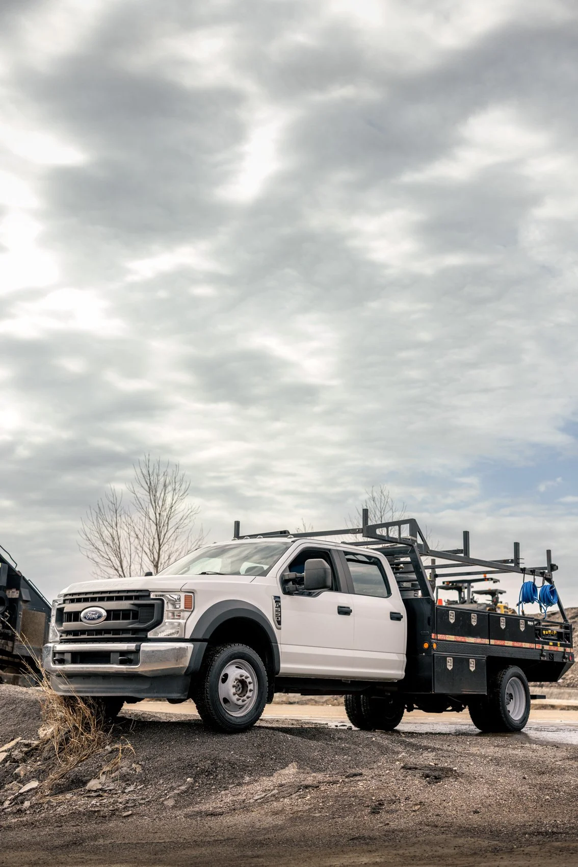 White Ford utility truck with a flatbed carrying equipment, parked on a gravel surface with cloudy sky and leafless trees in the background.