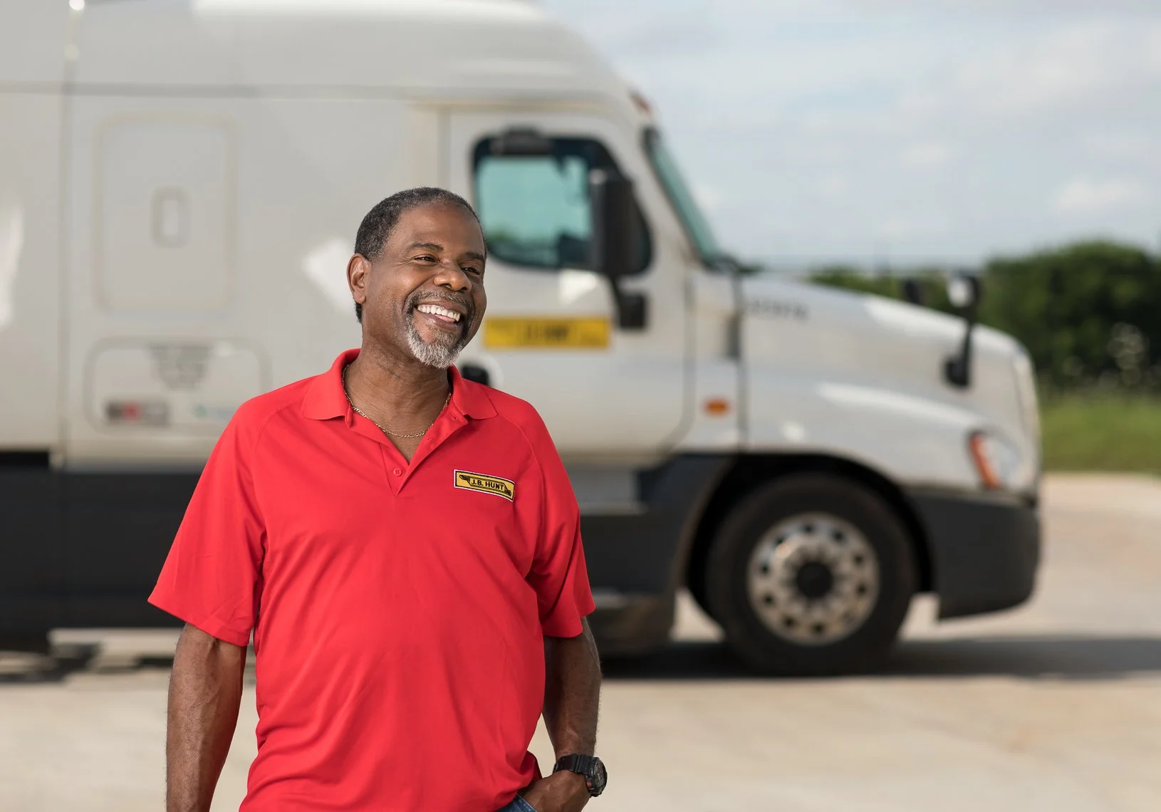 A smiling man in a red polo shirt stands outdoors in front of a large white truck. The man has a gray beard and is wearing a wristwatch, with a background of blue sky and greenery.